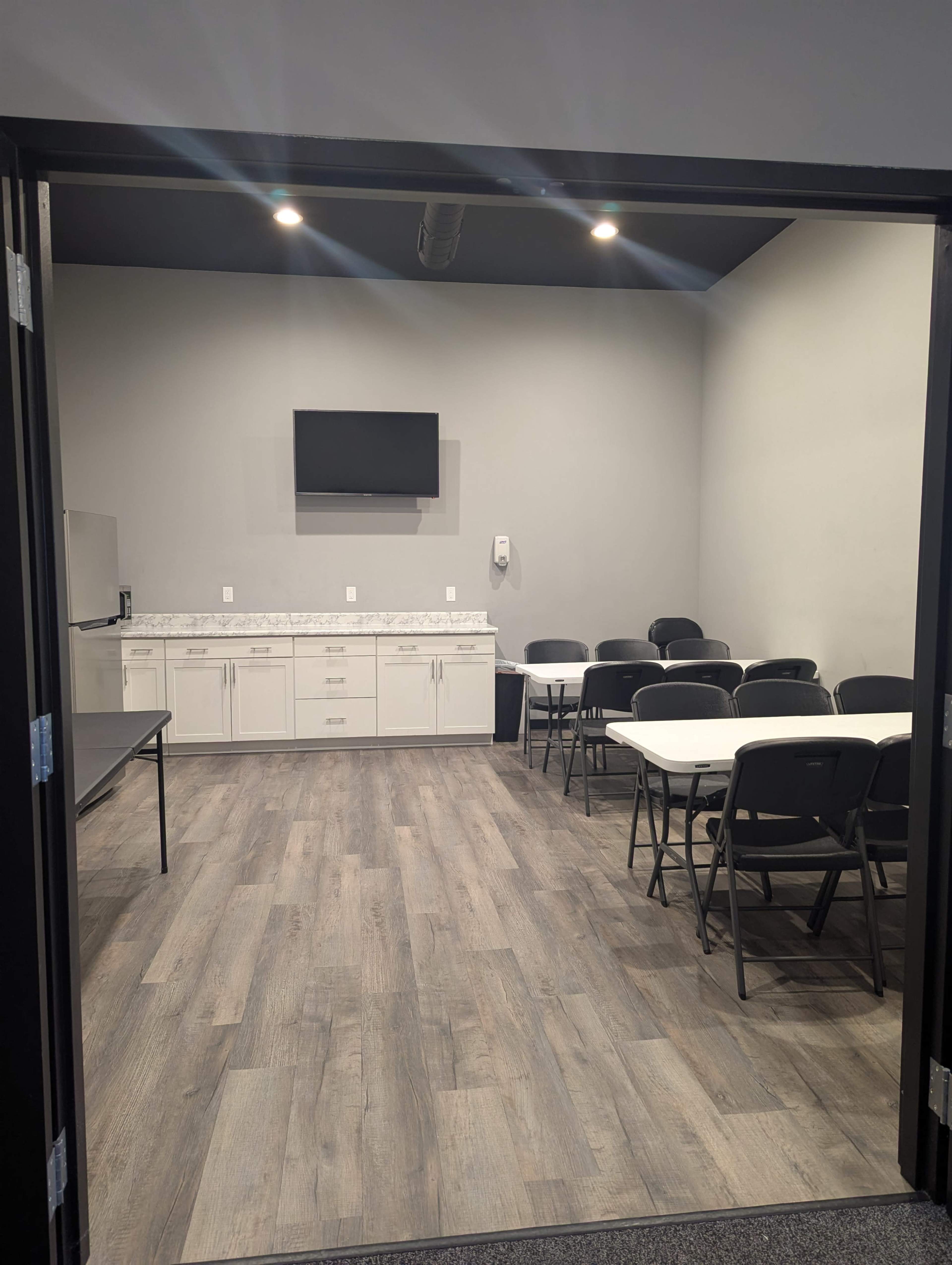 The image shows a meeting room with a large table, several chairs, a kitchenette area, and a wall-mounted television.
