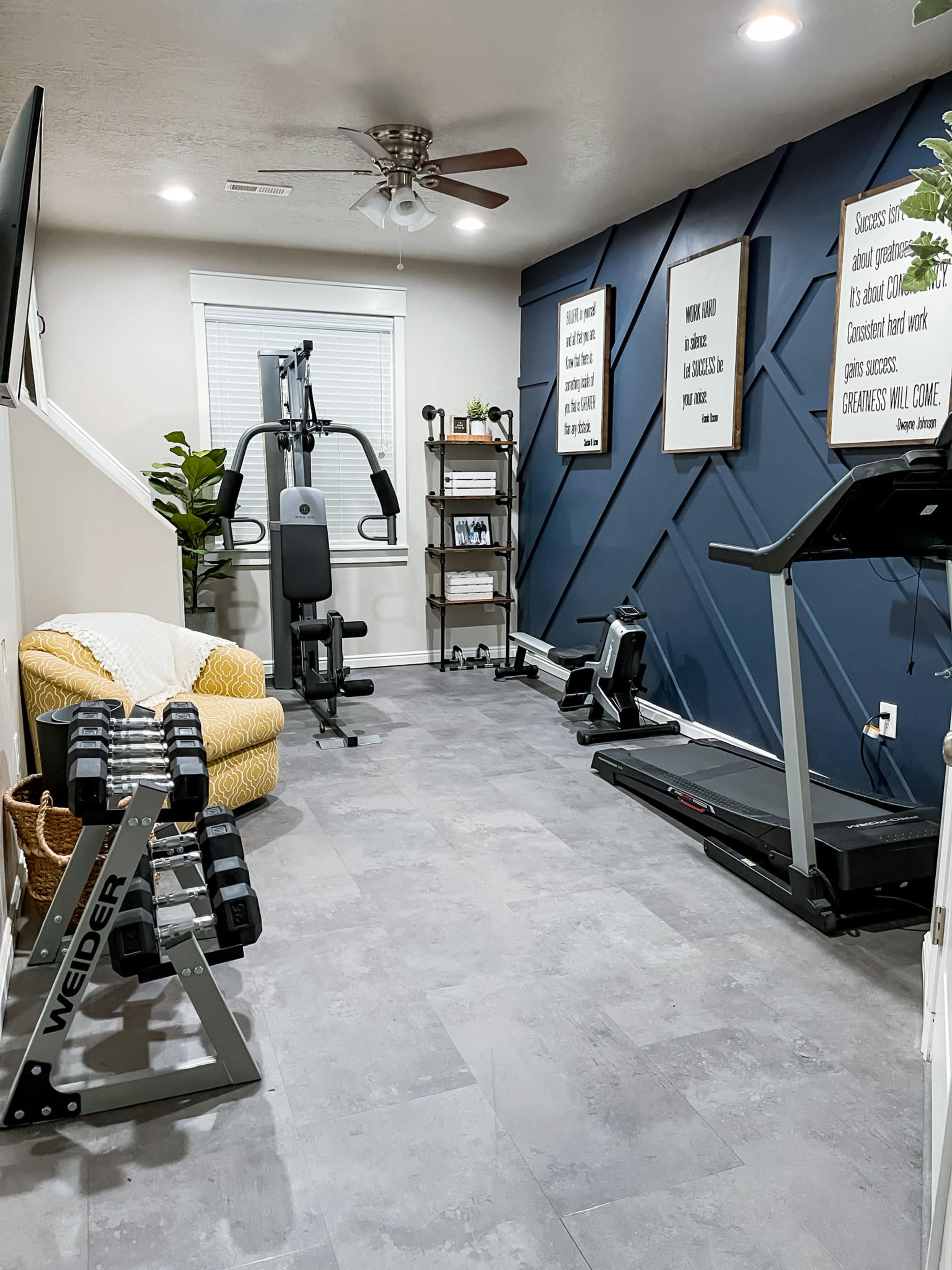 The image shows a home gym with exercise equipment, including a multi-gym machine, treadmill, and free weights, set against a blue accent wall and decorated with framed quotes.