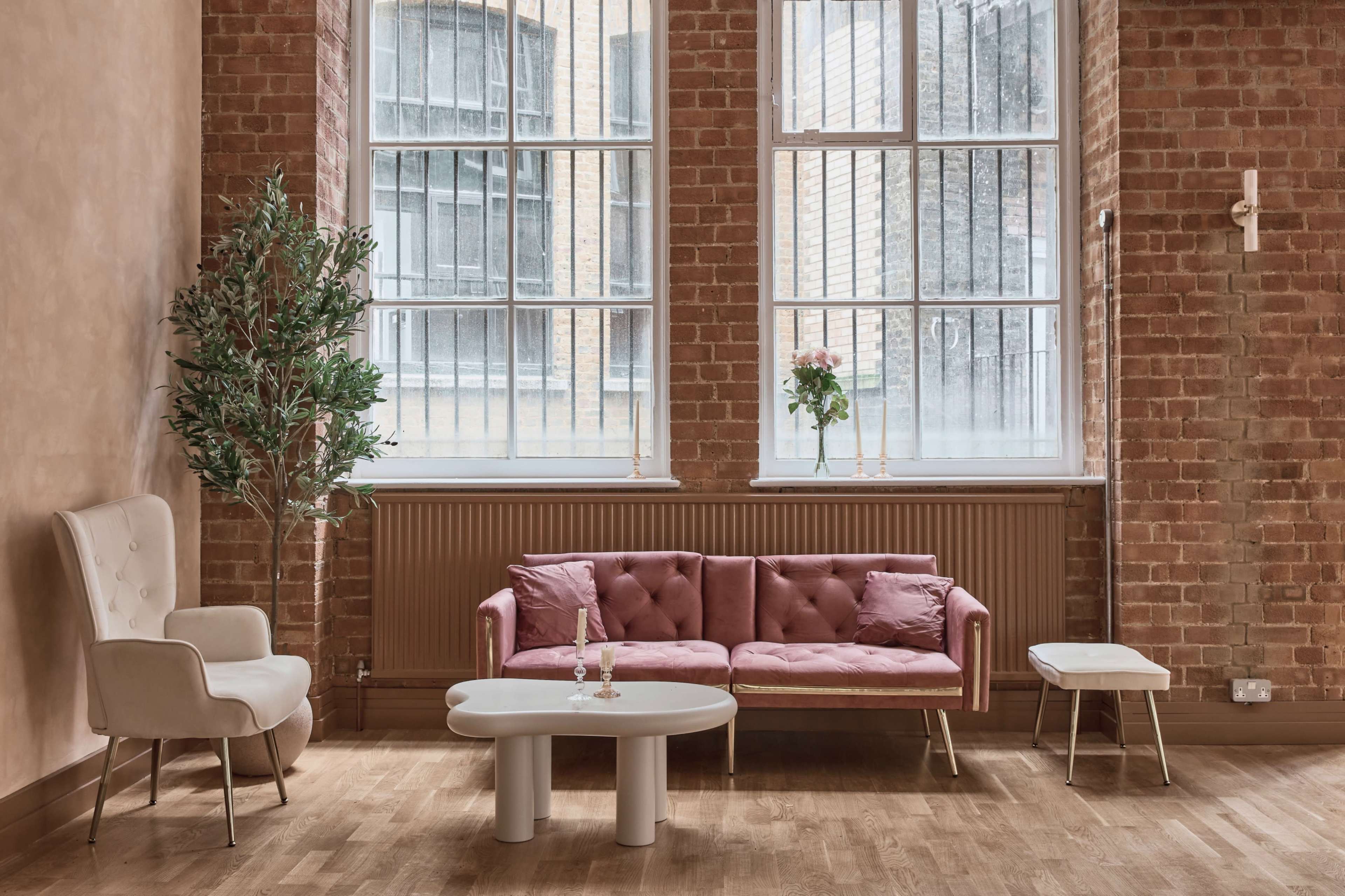 The image shows a modern living space with large windows, a pink tufted sofa, a small white table, and a potted plant, all set against a brick wall.