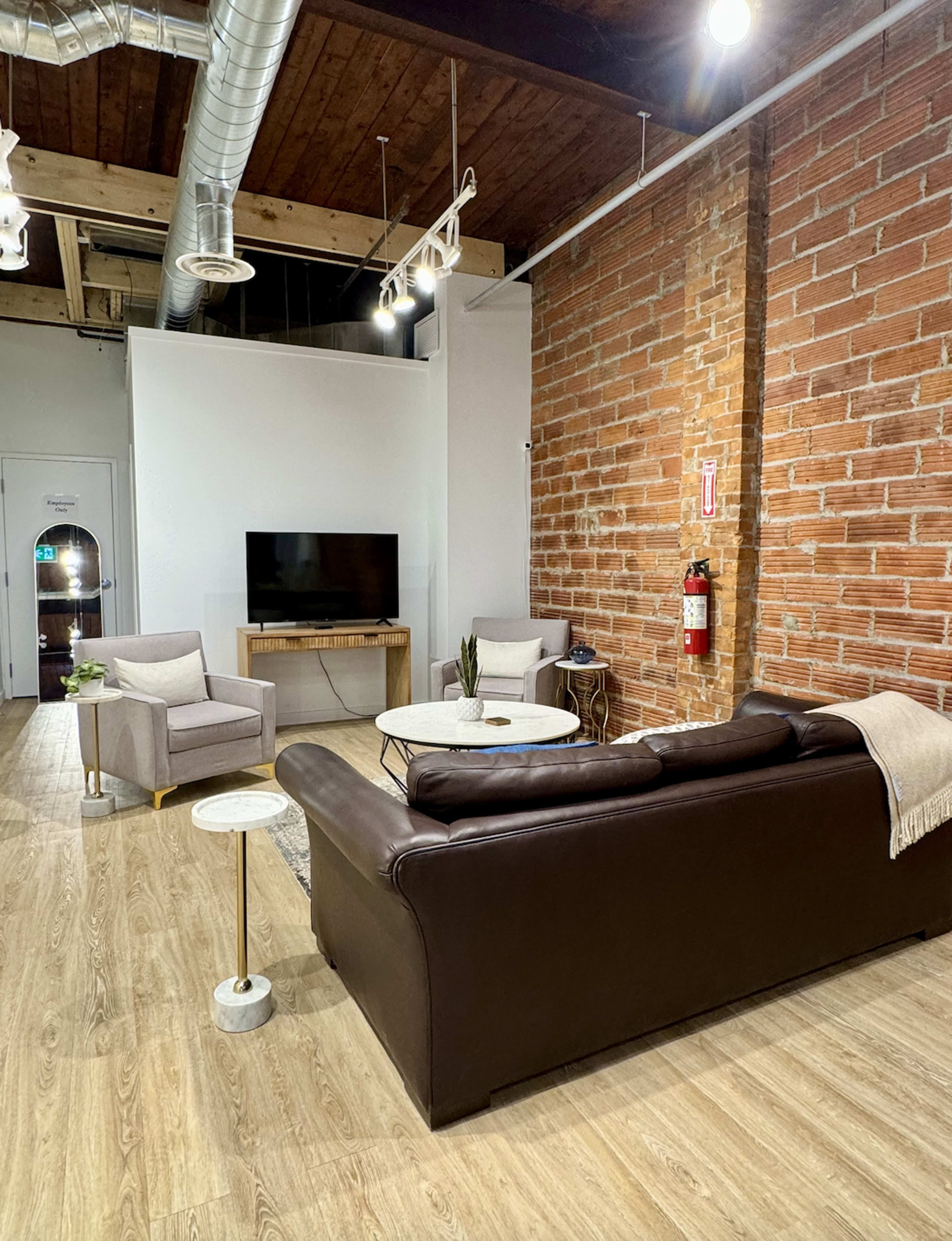 The image shows a modern living space with a brown leather sofa, a white coffee table, two armchairs, a television on a stand, and exposed brick walls.
