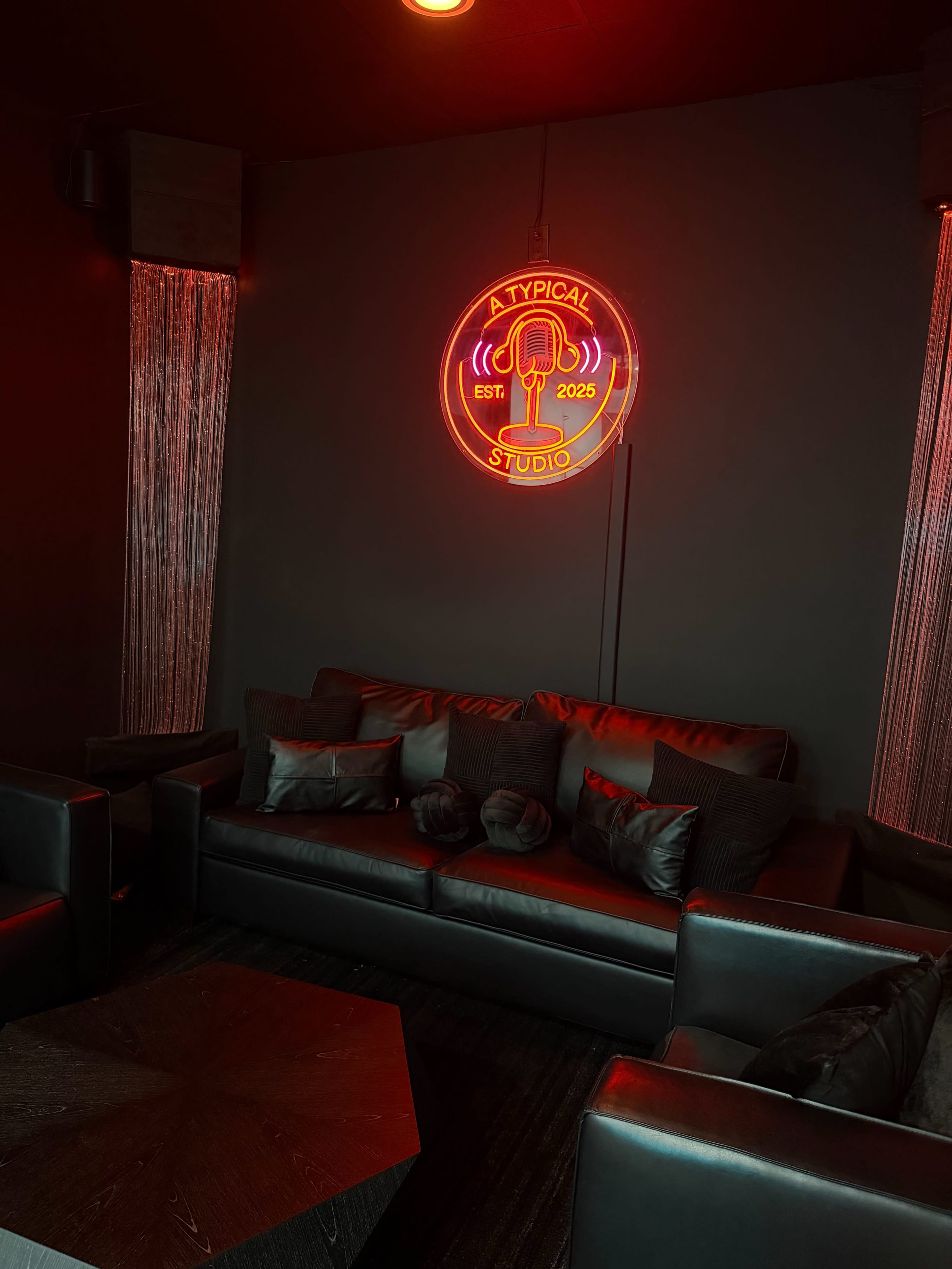 The image shows a dimly lit lounge area with black leather furniture and a neon sign that reads "A TYPICAL STUDIO" featuring a microphone motif.