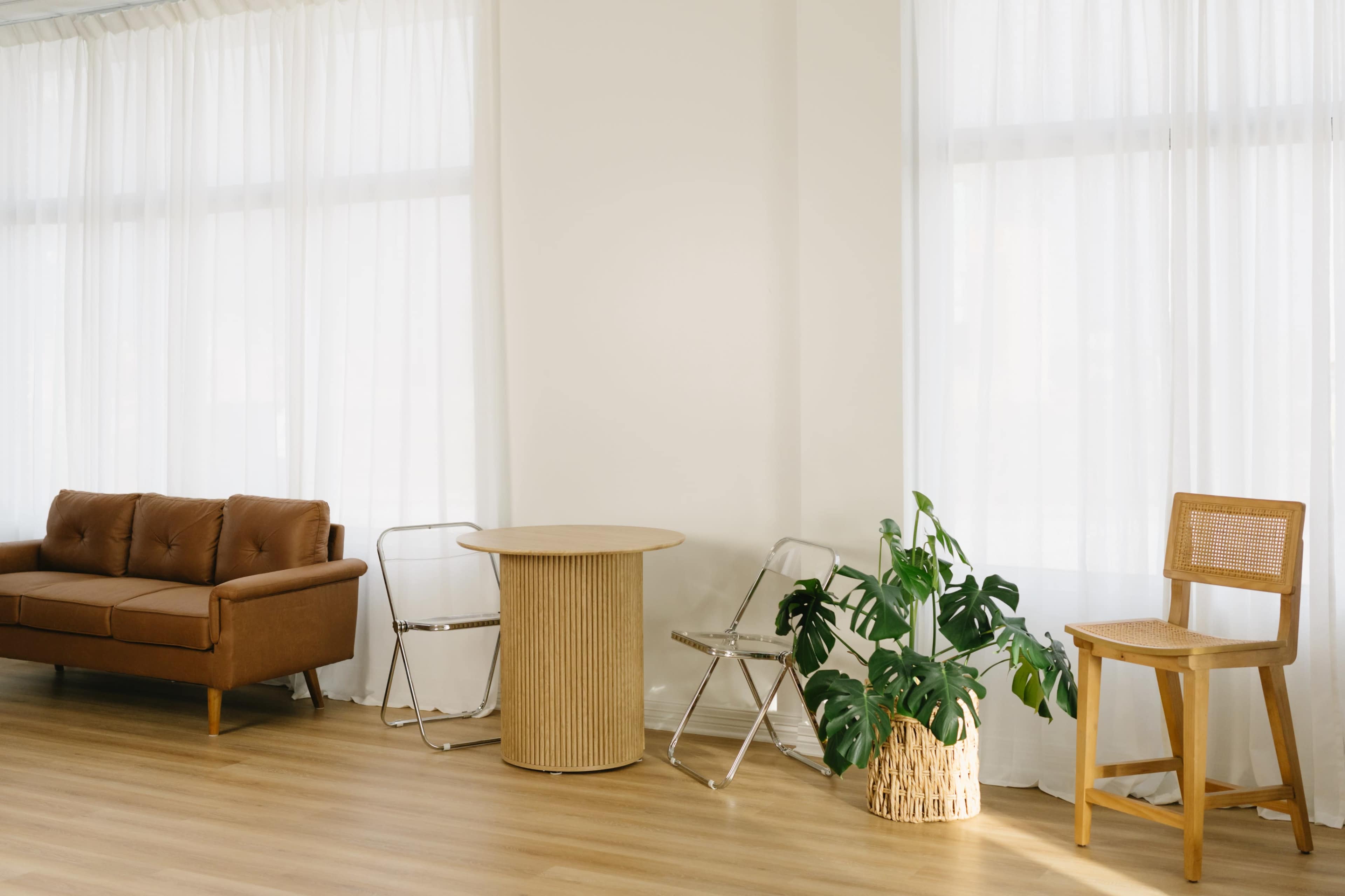 The image shows a light-filled interior space featuring a brown leather sofa, a round table, two metal chairs, a wooden chair, and a potted plant.