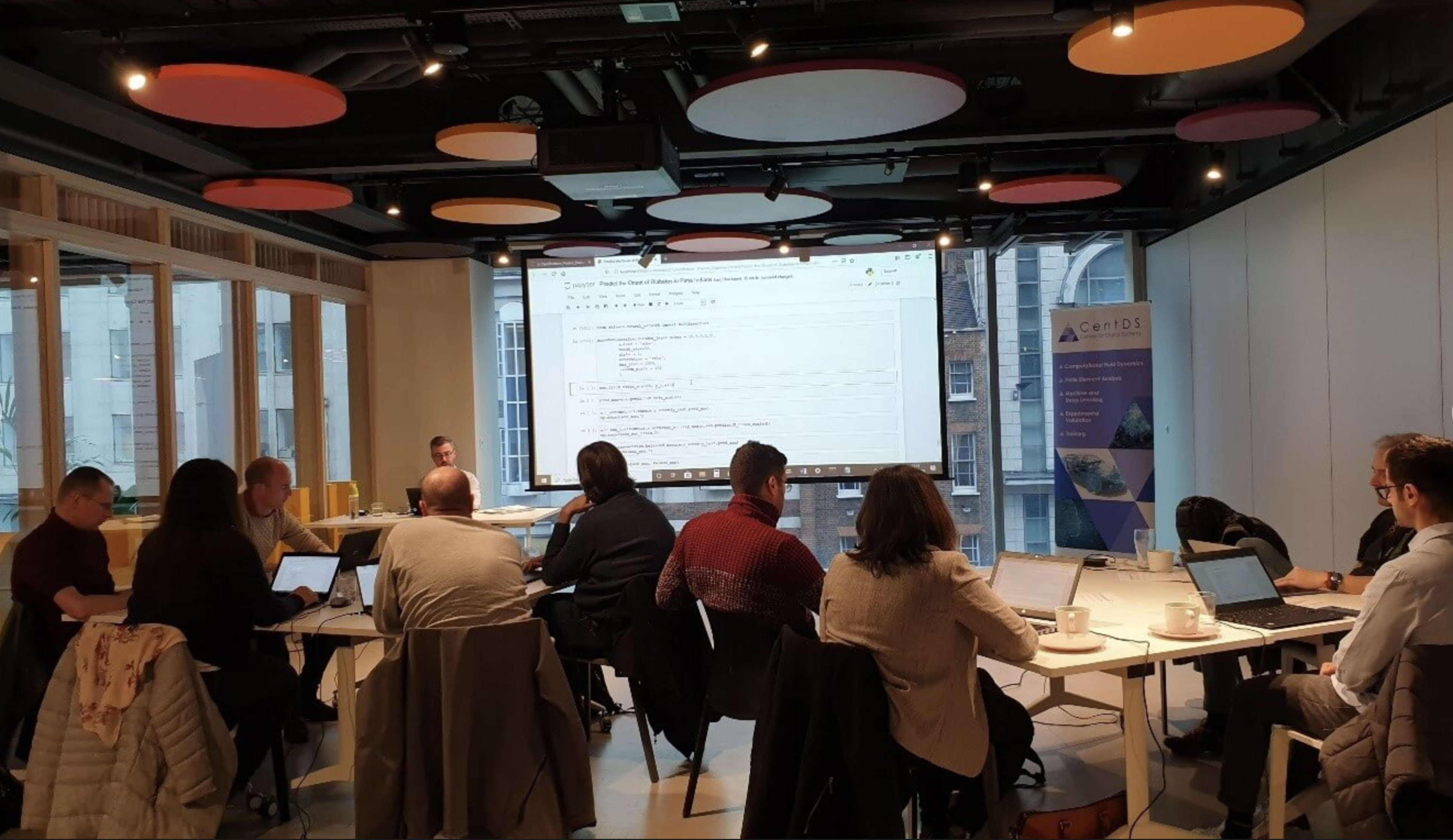 A group of participants is engaged in a training session around tables in a bright conference room, focusing on a presentation projected on a large screen.