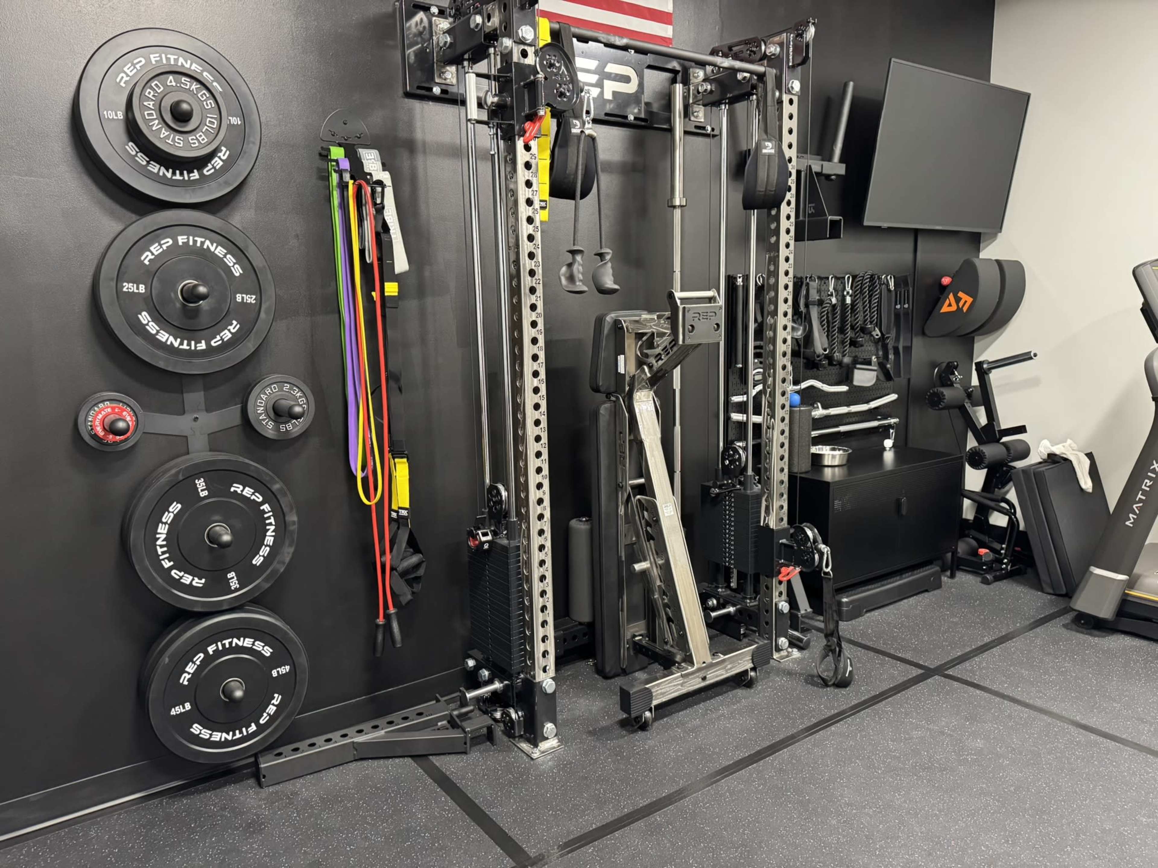 The image shows a home gym setup featuring a cable machine, weight plates mounted on the wall, resistance bands, and various exercise equipment in a dark-colored room.