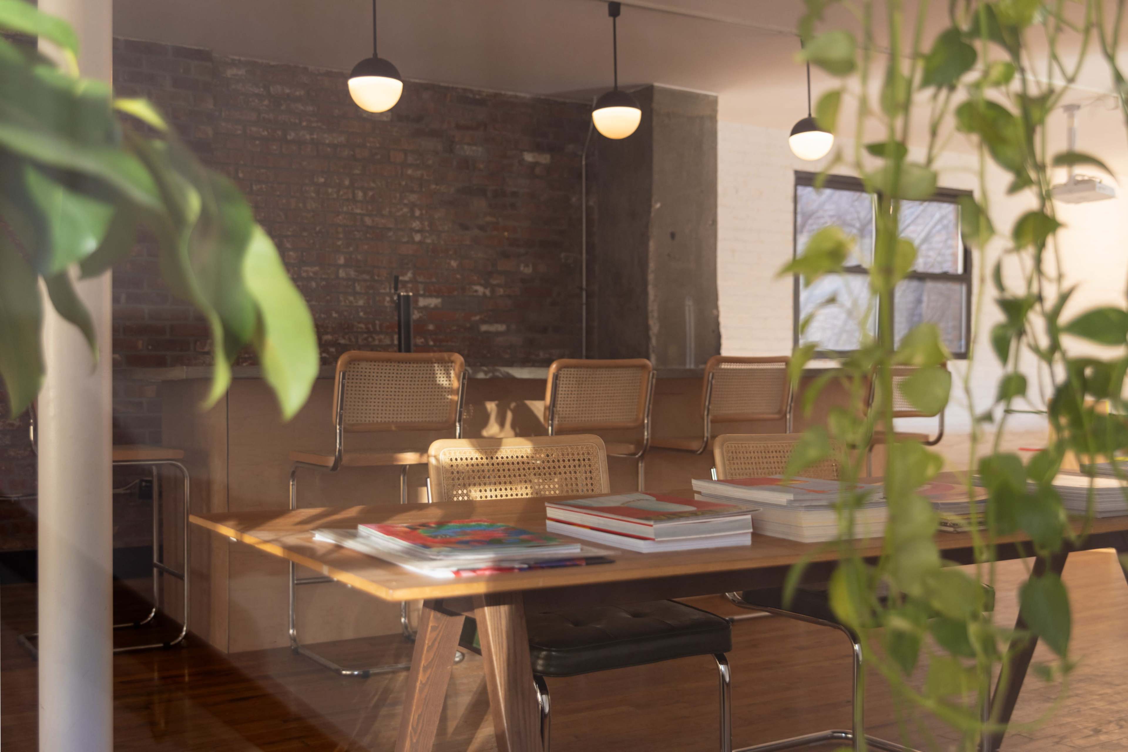 A modern meeting room with wooden furniture, stacks of magazines on a table, and plants framing the view through a glass wall.