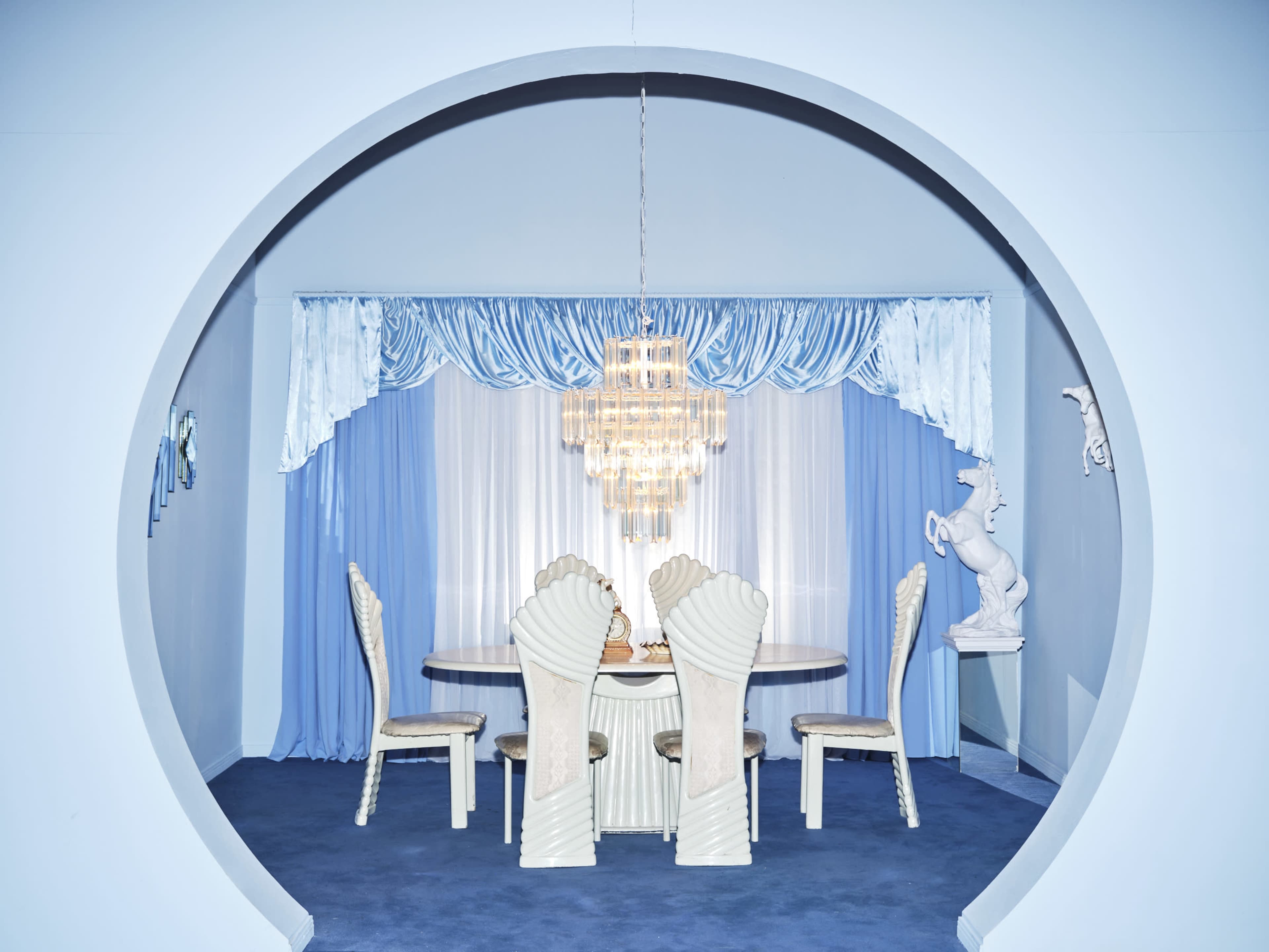 A circular arch frames a dining area with a round table and chairs, illuminated by a chandelier, surrounded by light blue curtains.