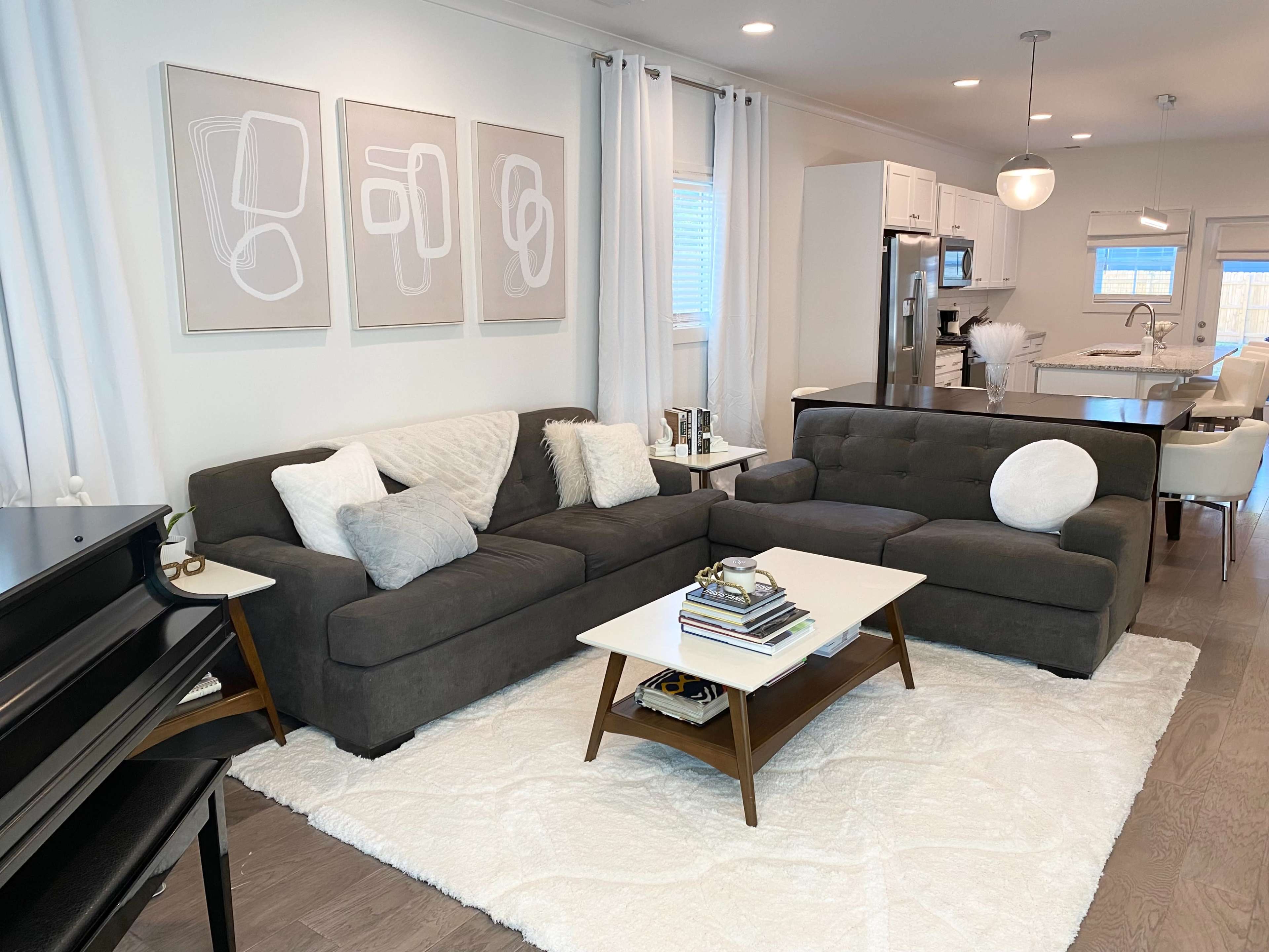 The image shows a modern living room featuring a dark sectional sofa, a white coffee table, and framed abstract artwork on the wall, with a kitchen visible in the background.