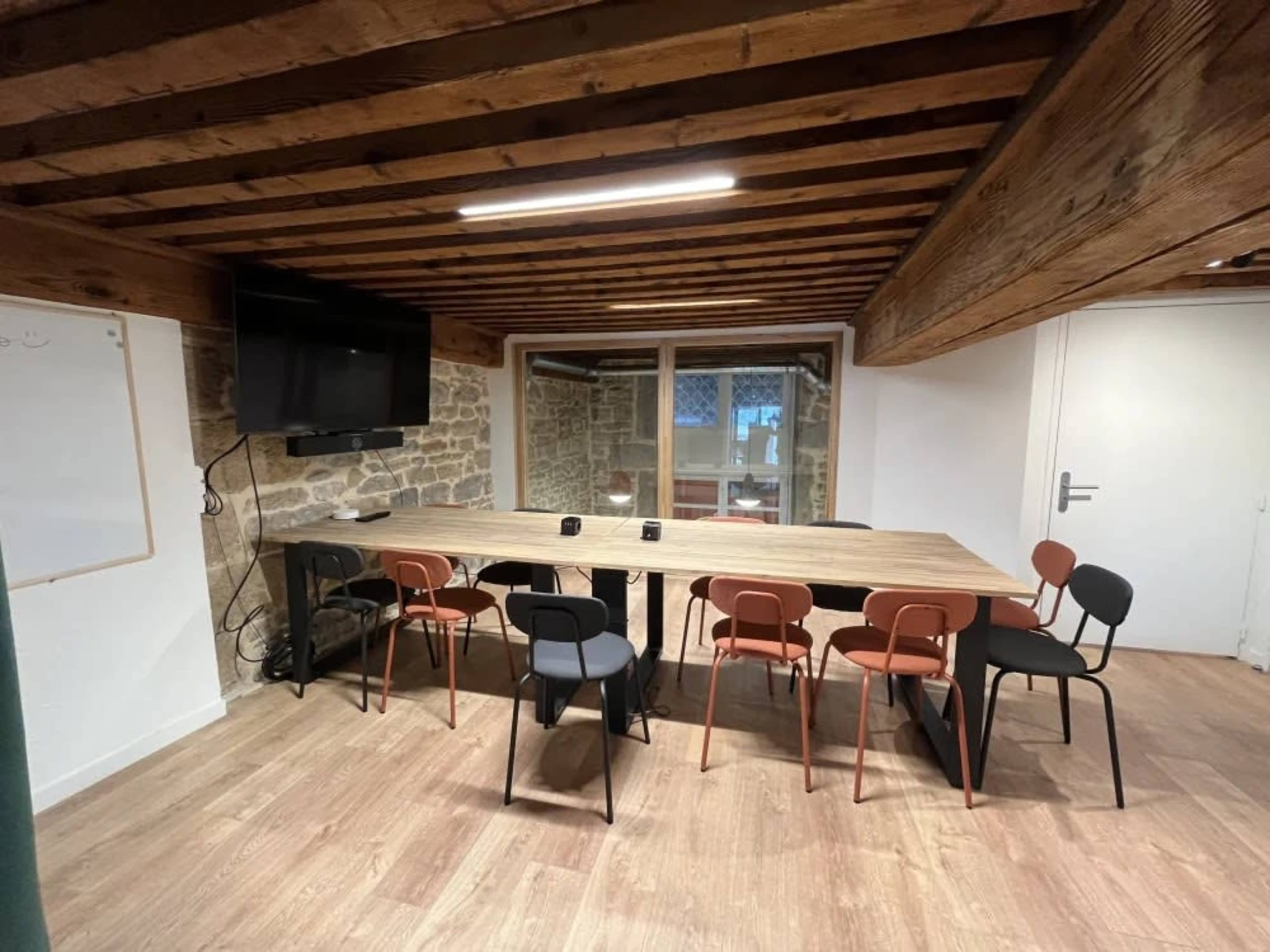 A modern meeting room features a long table surrounded by different colored chairs, with exposed wooden beams and a large window.
