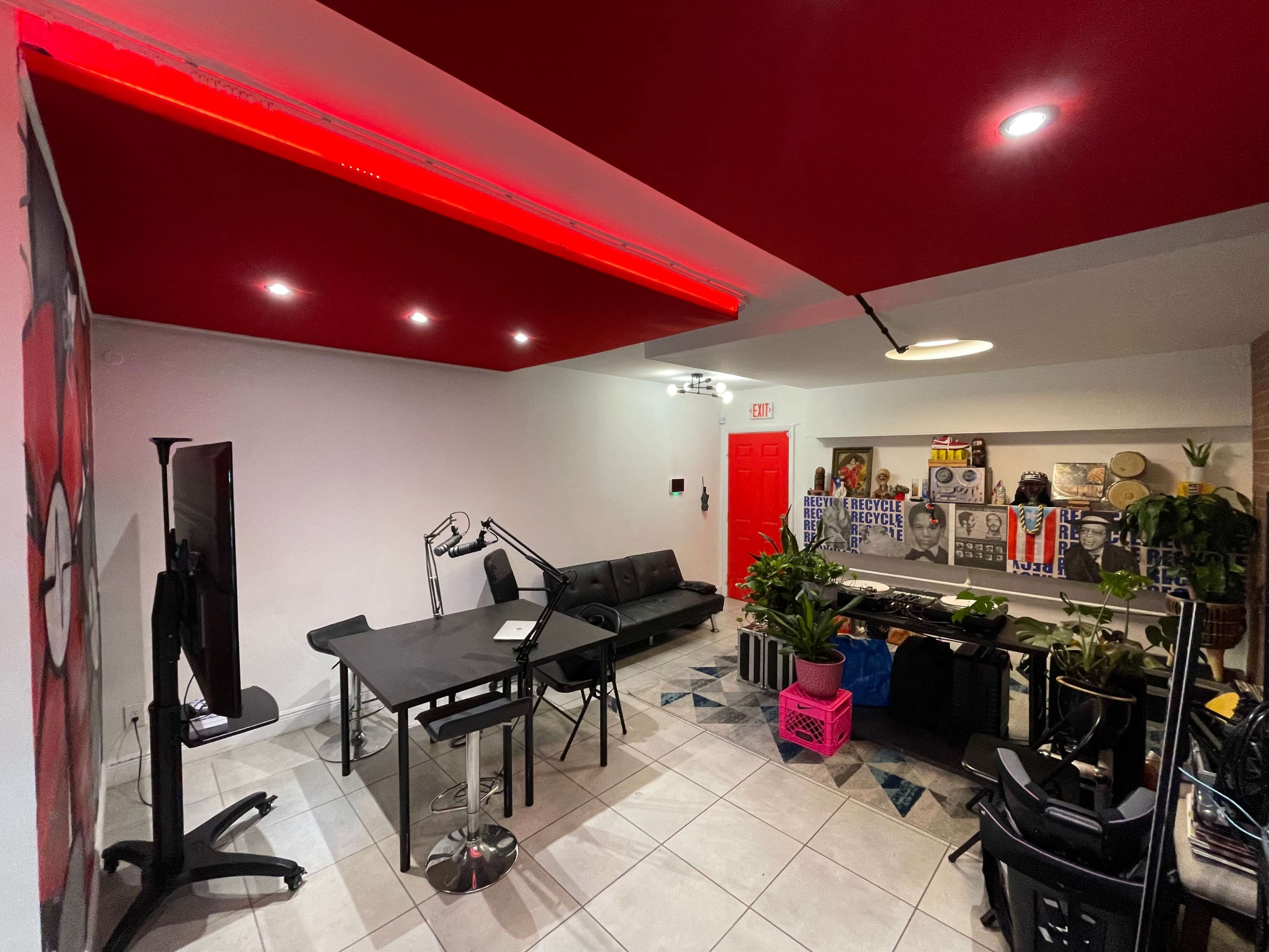 A modern studio space featuring a black sofa, a glass table, and various plants, with red lighting along the ceiling and a bright red door.