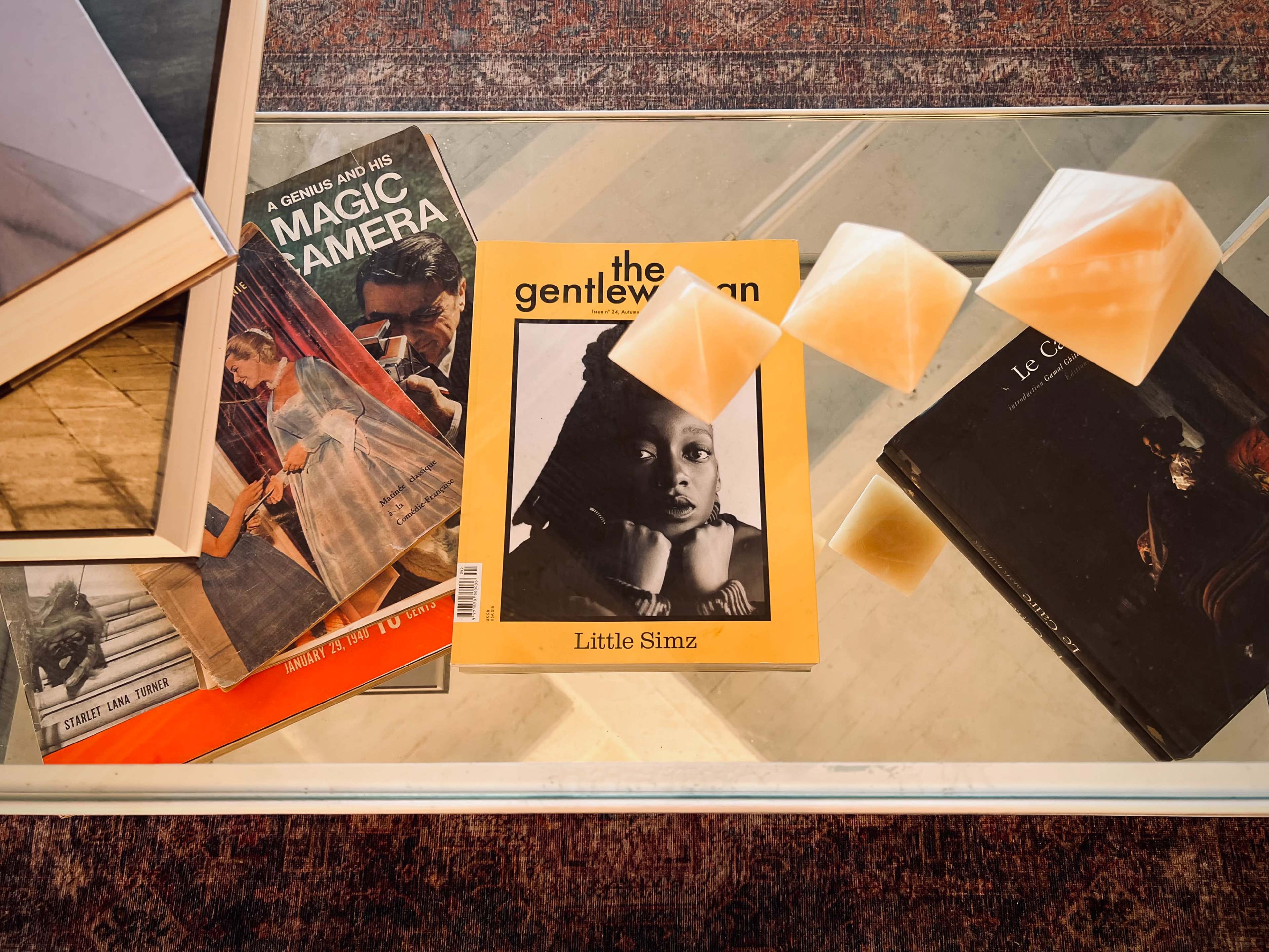 A glass table displays a collection of books, including "The Gentleman" featuring Little Simz, along with several soft, triangular-shaped objects.