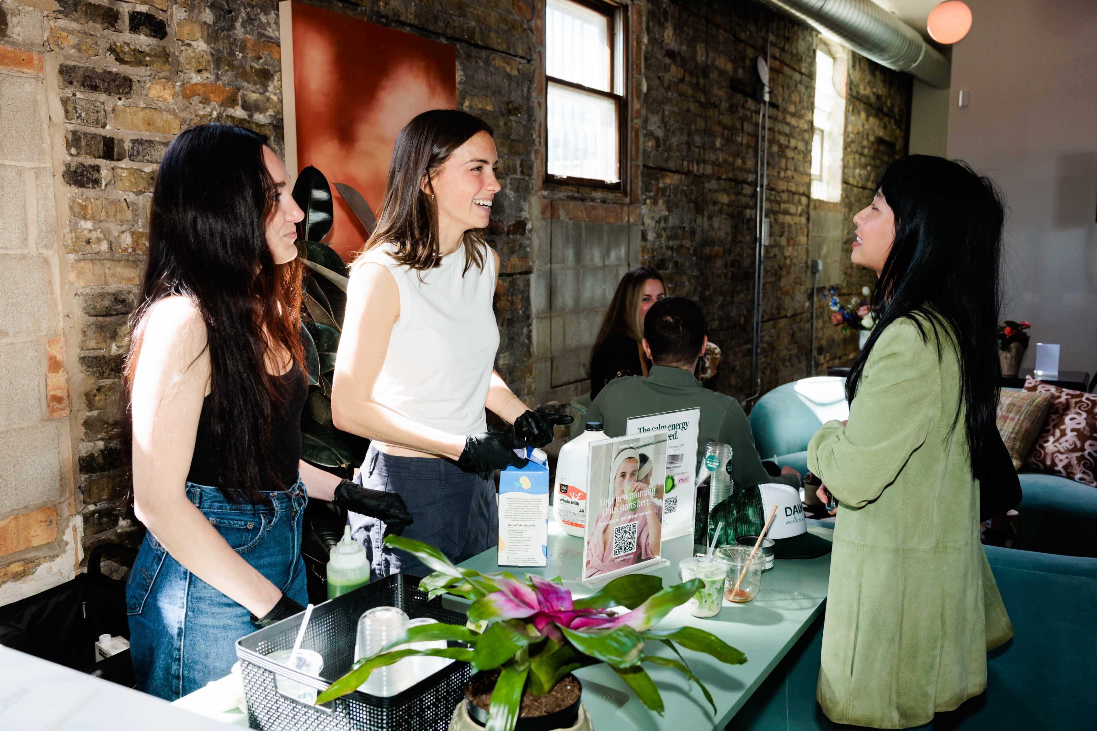 Three women are interacting in a stylish indoor space with exposed brick walls, one woman wearing gloves appears to be speaking to the others, while a fourth person is seated in the background.