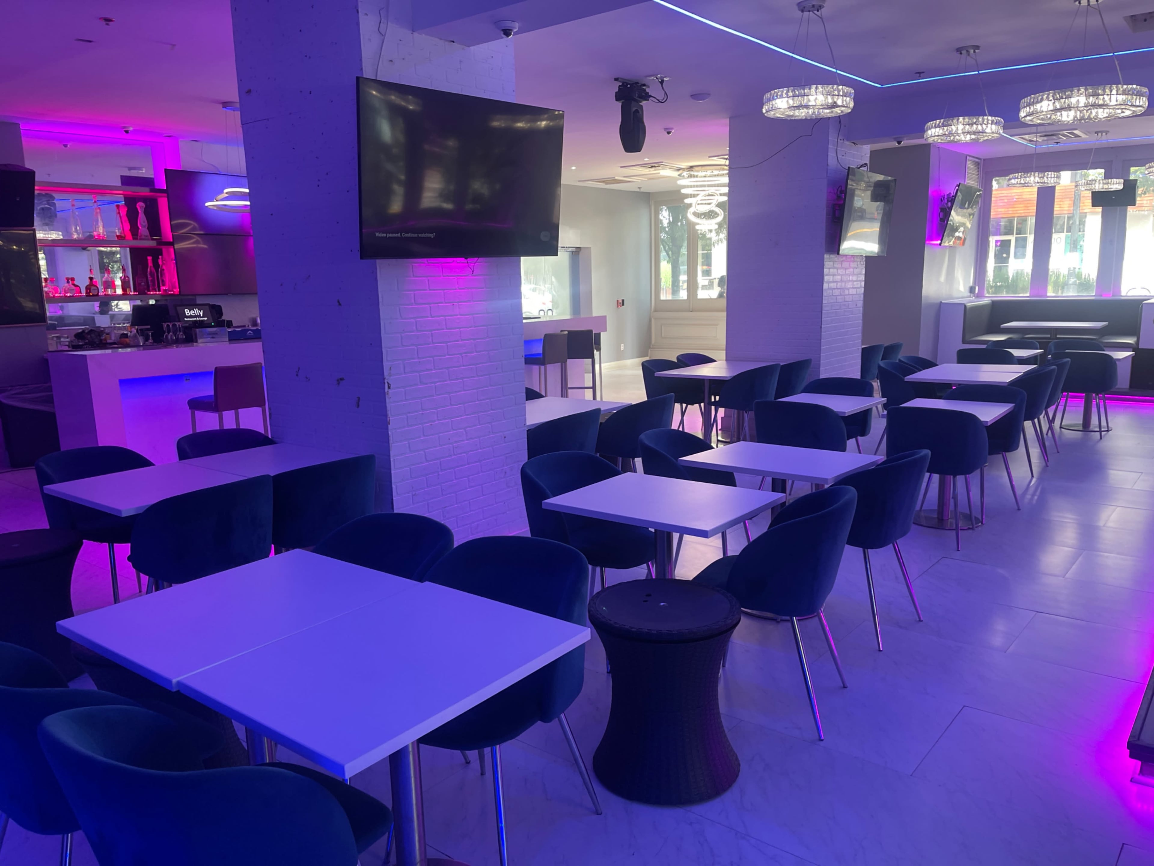 An empty dining area with blue chairs and white tables, illuminated by purple and white lighting, and a bar in the background.