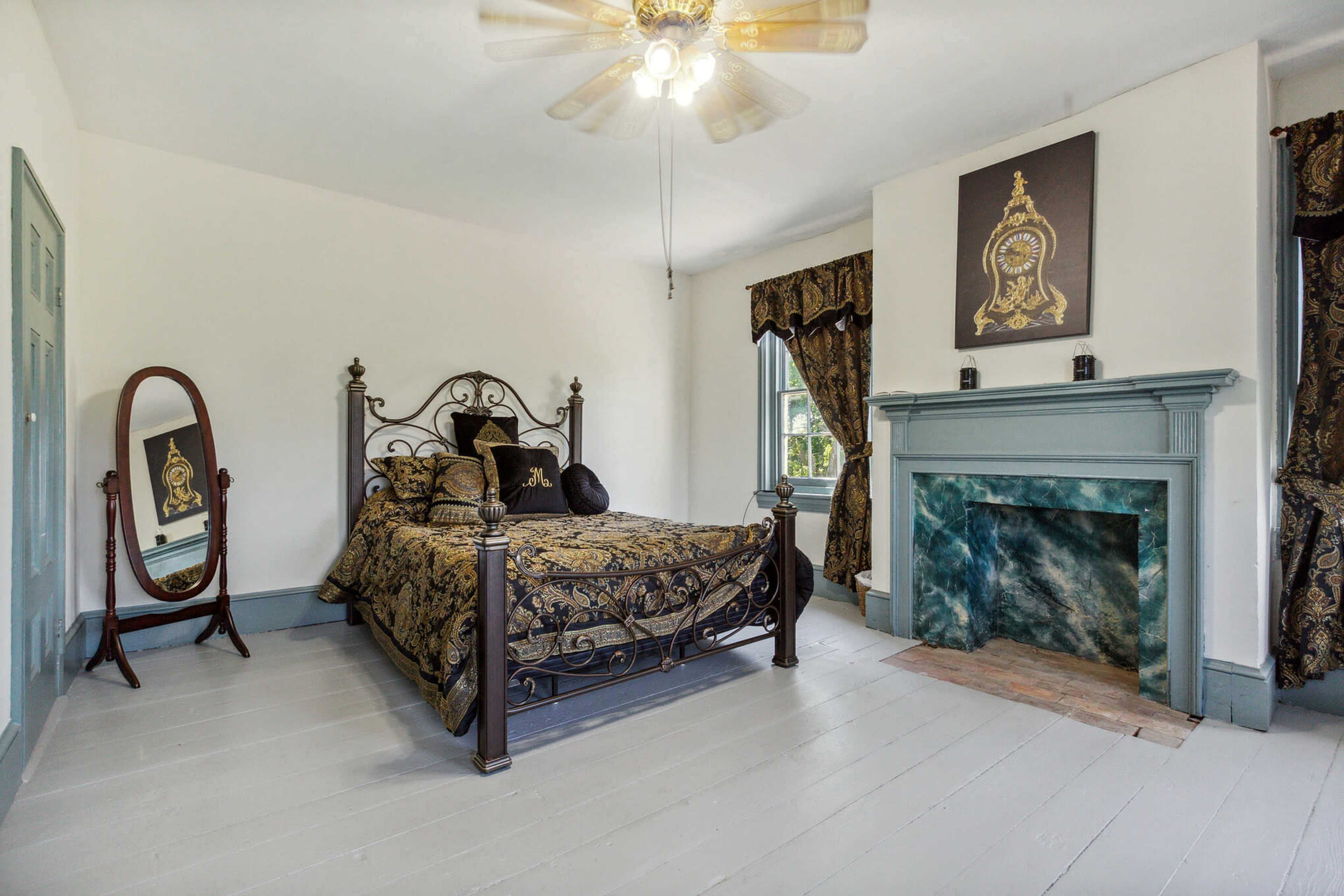 The image shows a bedroom featuring a wrought iron bed, a large mirror, a fireplace with a blue-green mantle, and patterned curtains.