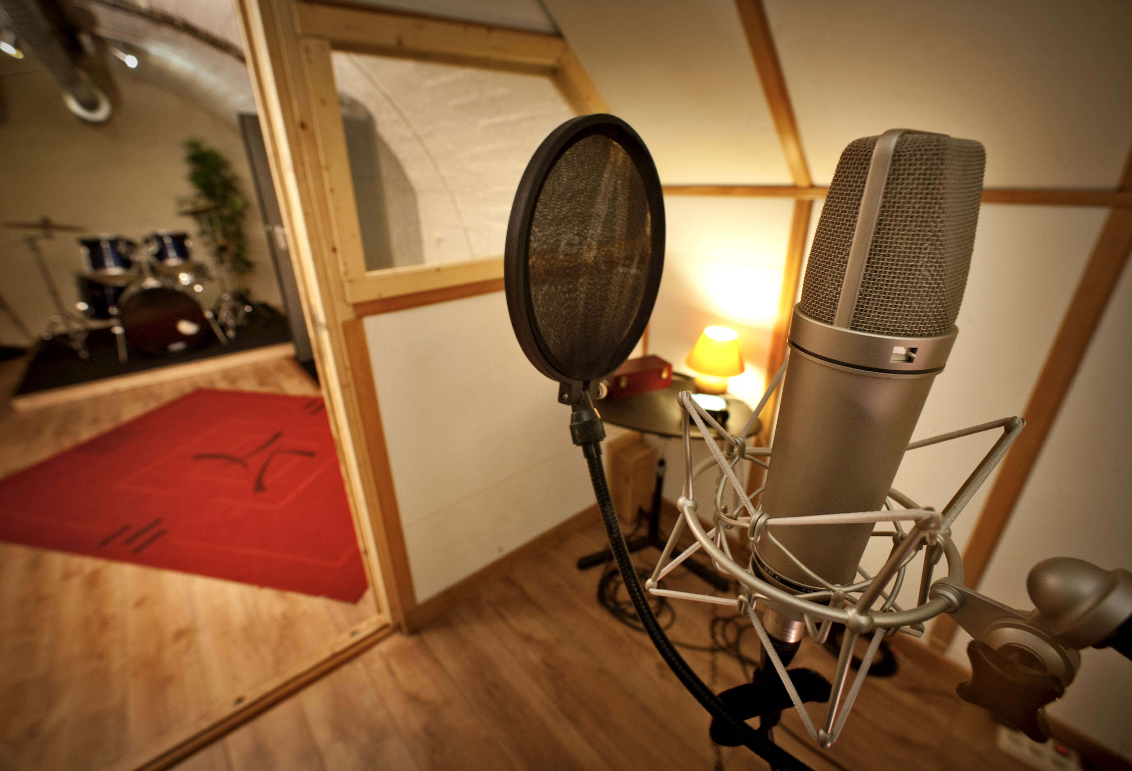 A microphone stands in the foreground with a soundproof booth and a drum set visible in the background.