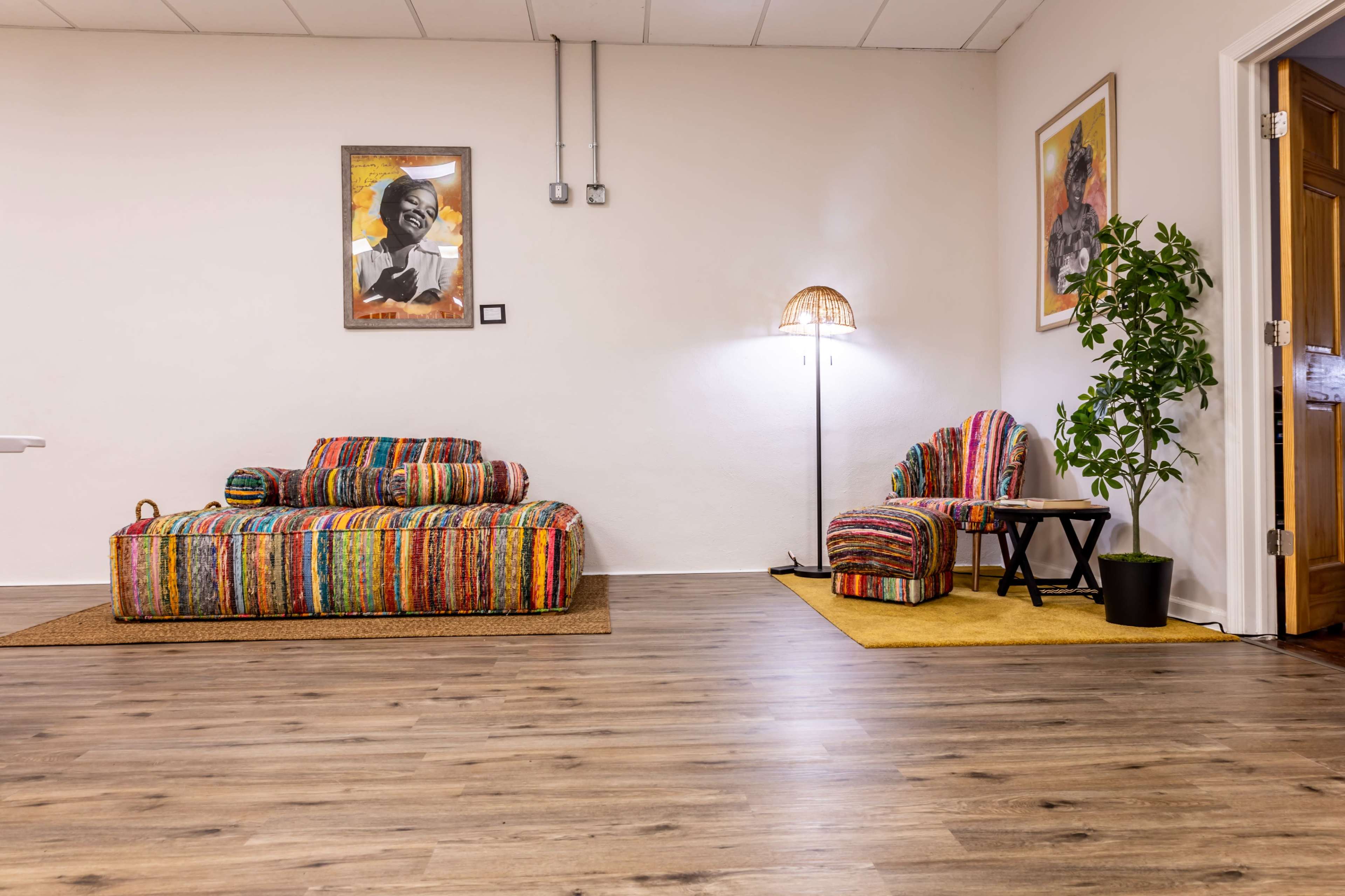 The image shows a room with colorful, patterned seating, a lamp, a small table, a potted plant, and framed artwork on the walls.