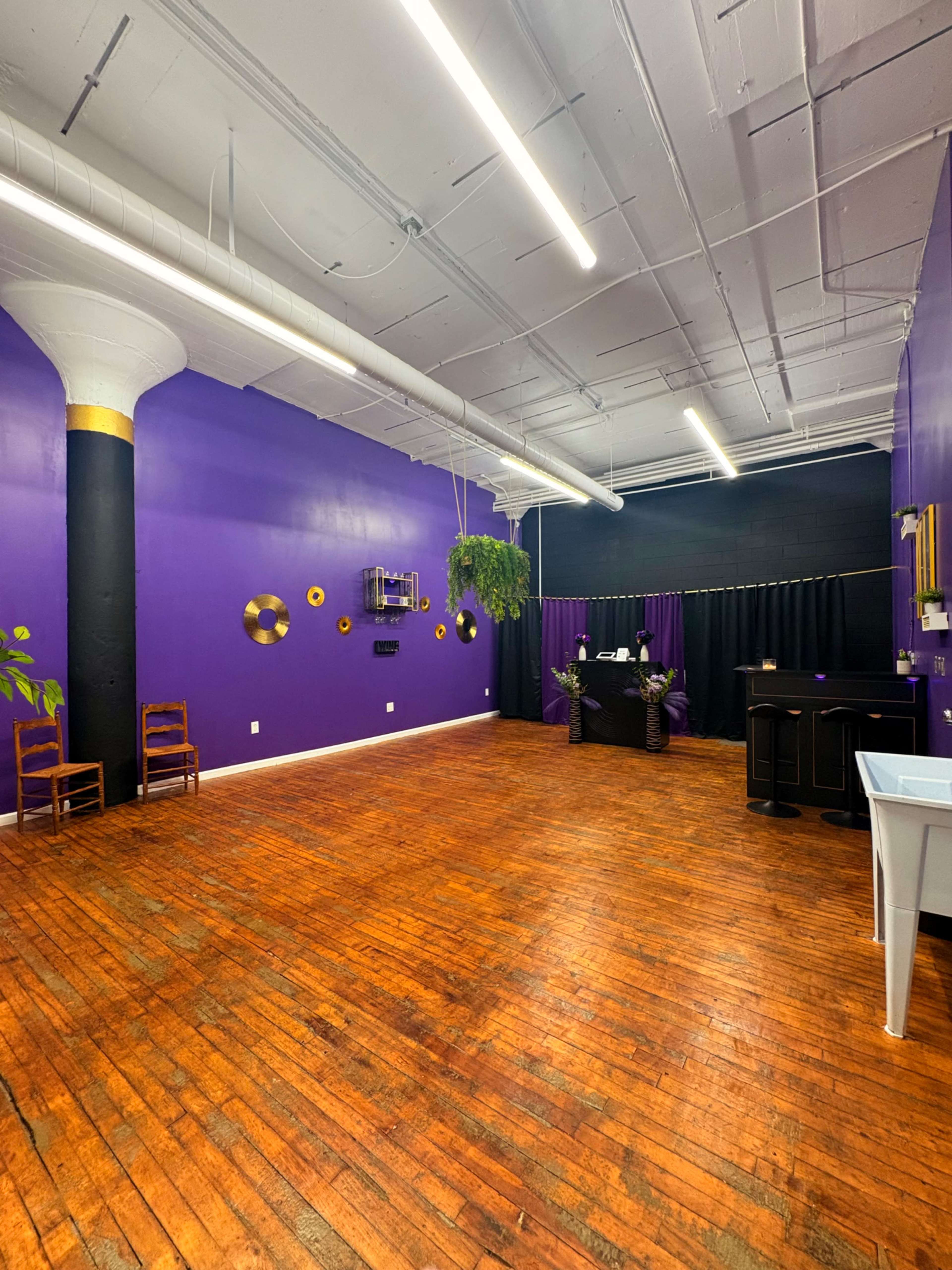 The image shows an empty room with purple walls, wooden floors, and a reception area featuring a desk and decorative elements.