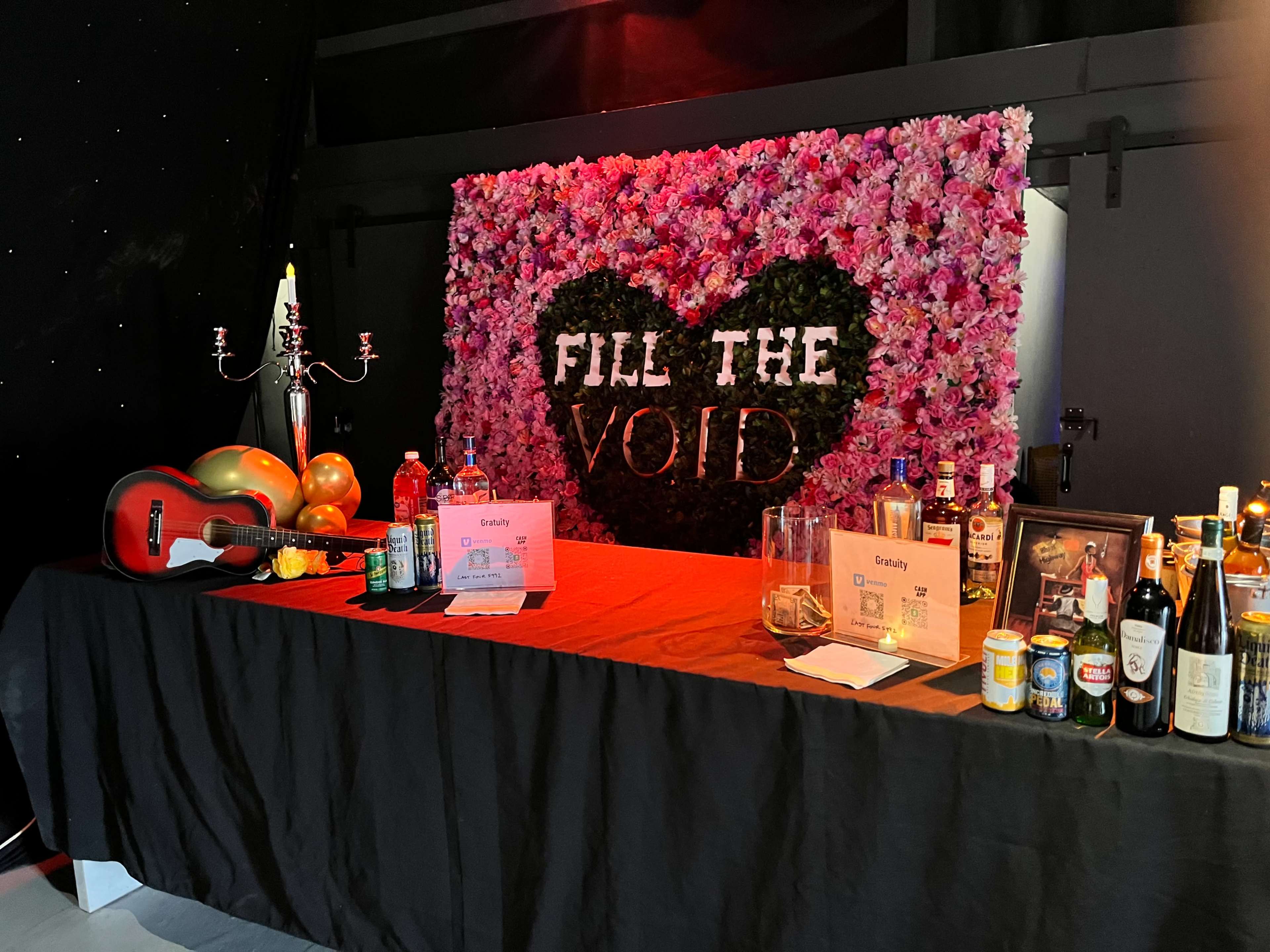 A bar setup features a heart-shaped floral display with the words "FILL THE VOID" and a variety of drinks and decorations on the table.