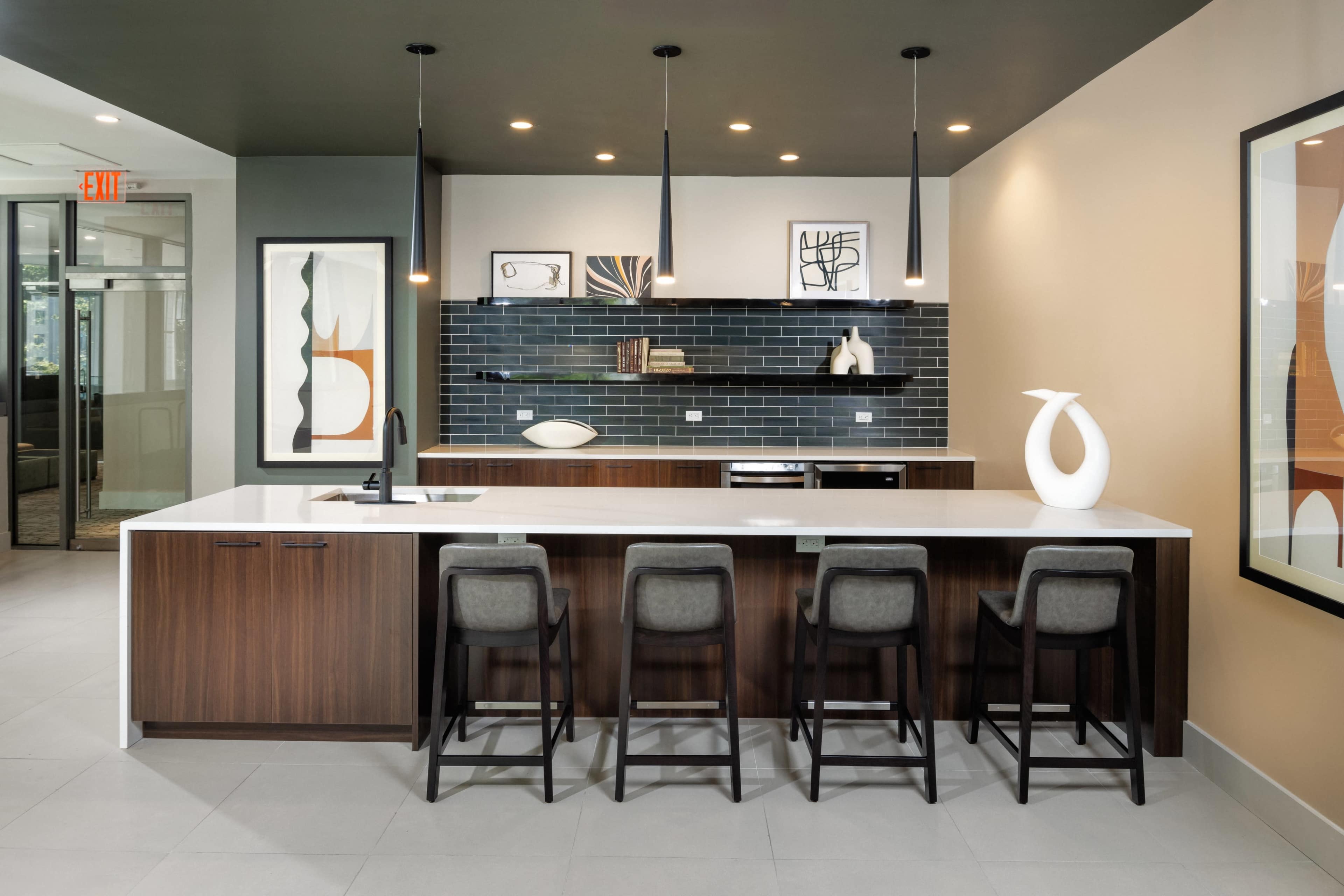 The image shows a modern kitchen area featuring a dark wooden counter with bar stools, a white countertop, and a backsplash made of dark tiles.