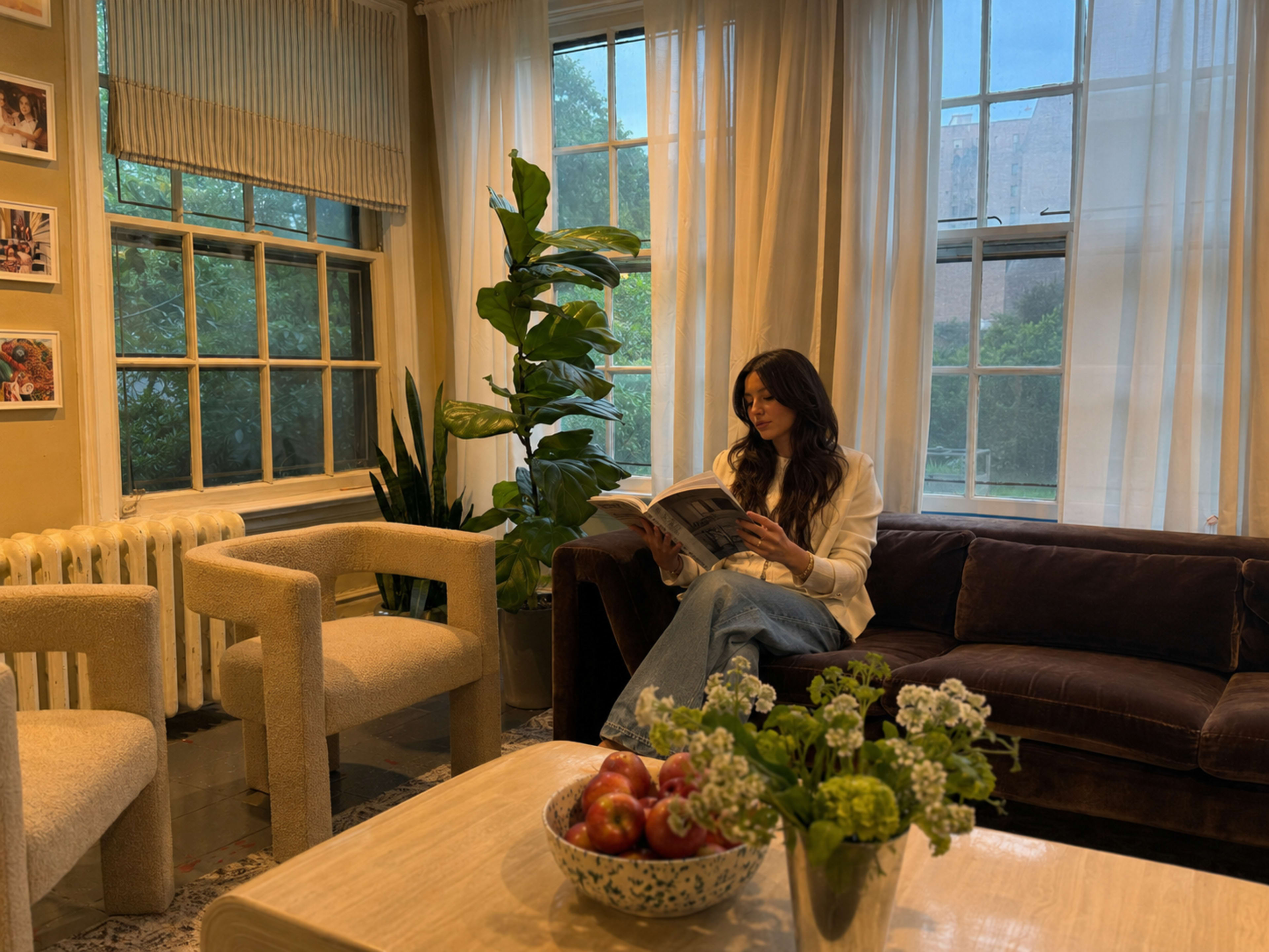 A woman reading a magazine sits on a brown sofa in a cozy room with large windows and potted plants.