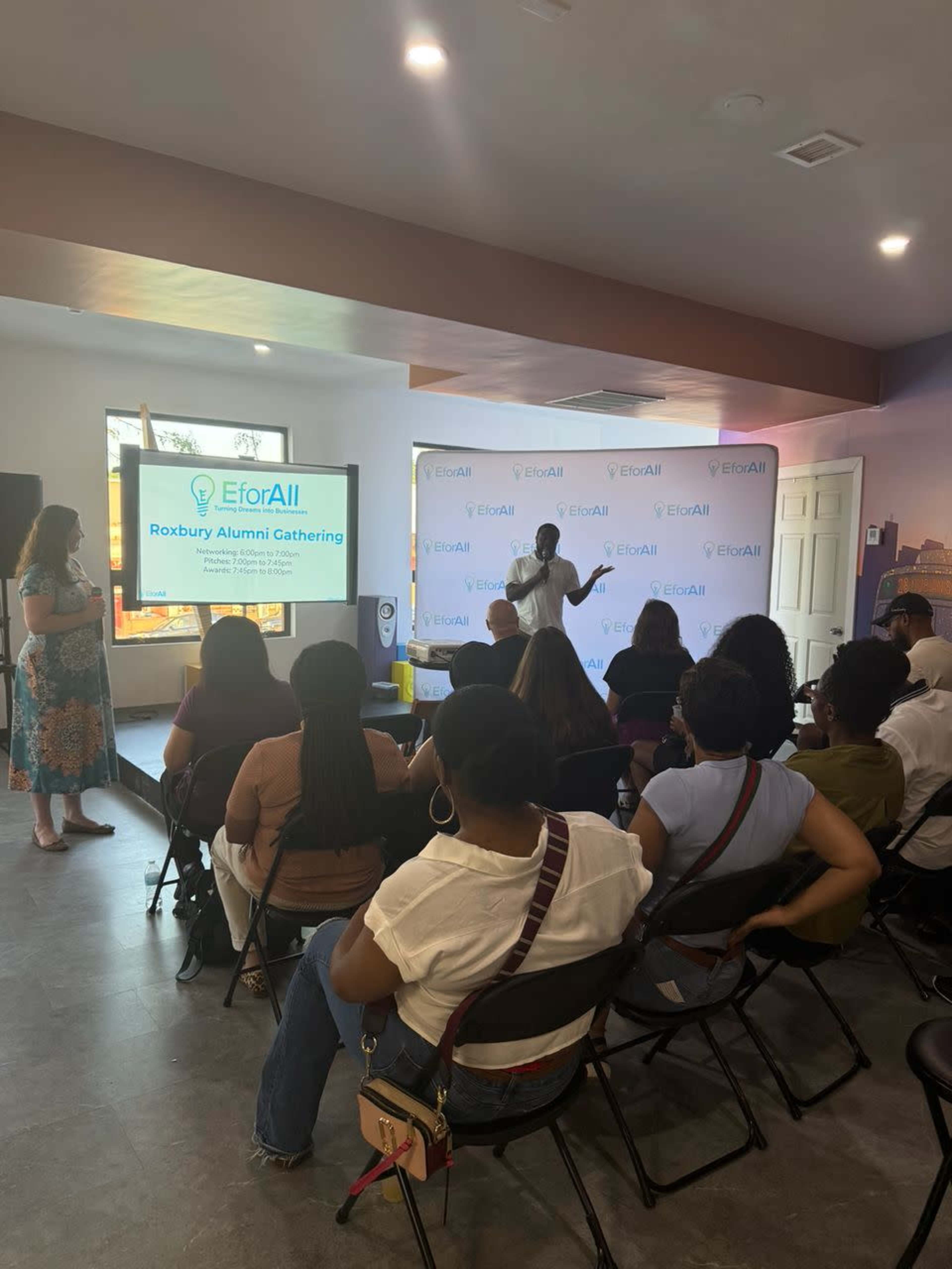 A speaker presents at the EforAll Roxbury Alumni Gathering in a well-lit room with an audience seated in rows.