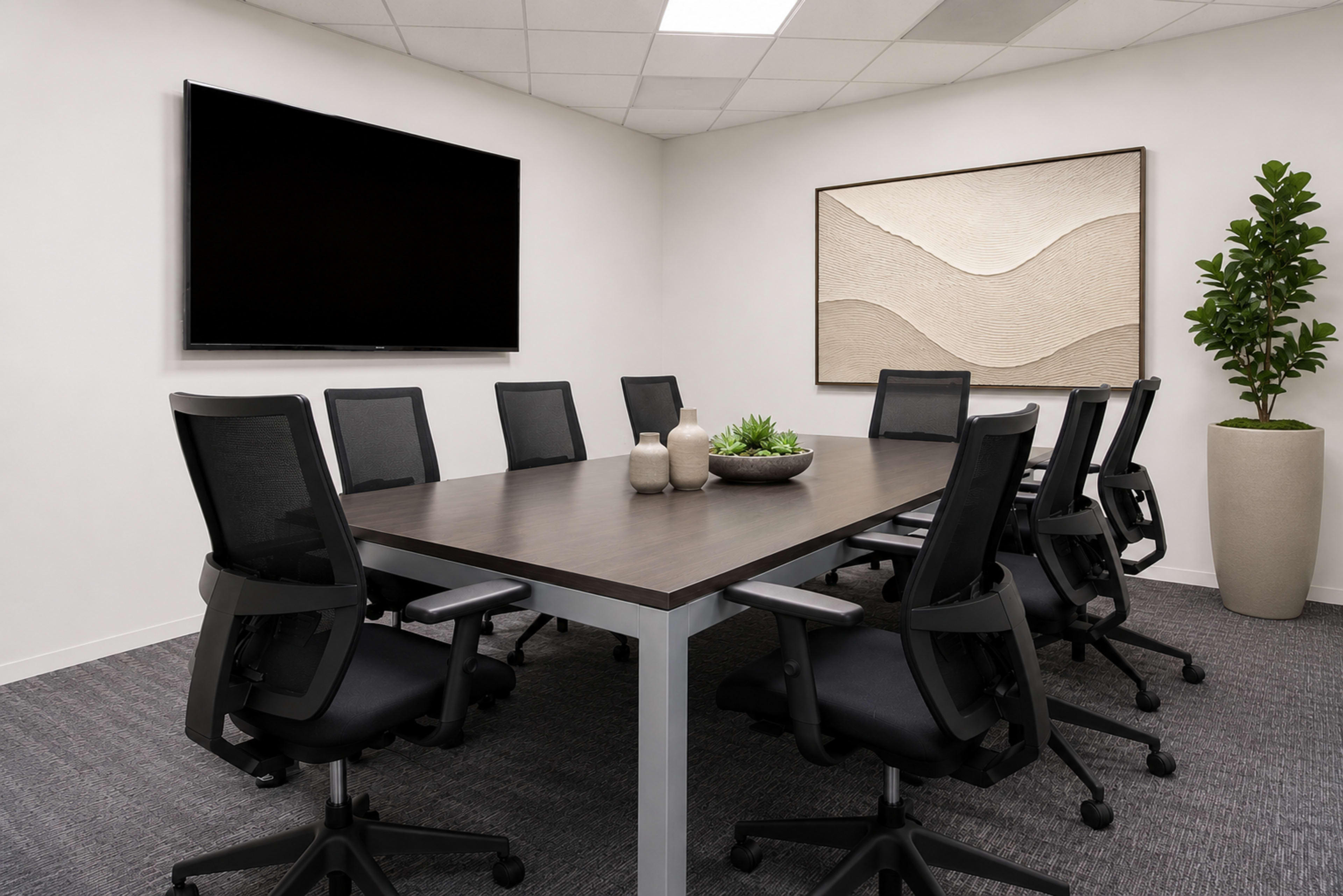 A modern conference room features a long table surrounded by ten black chairs, a large screen on one wall, and abstract artwork along with a decorative plant in the corner.