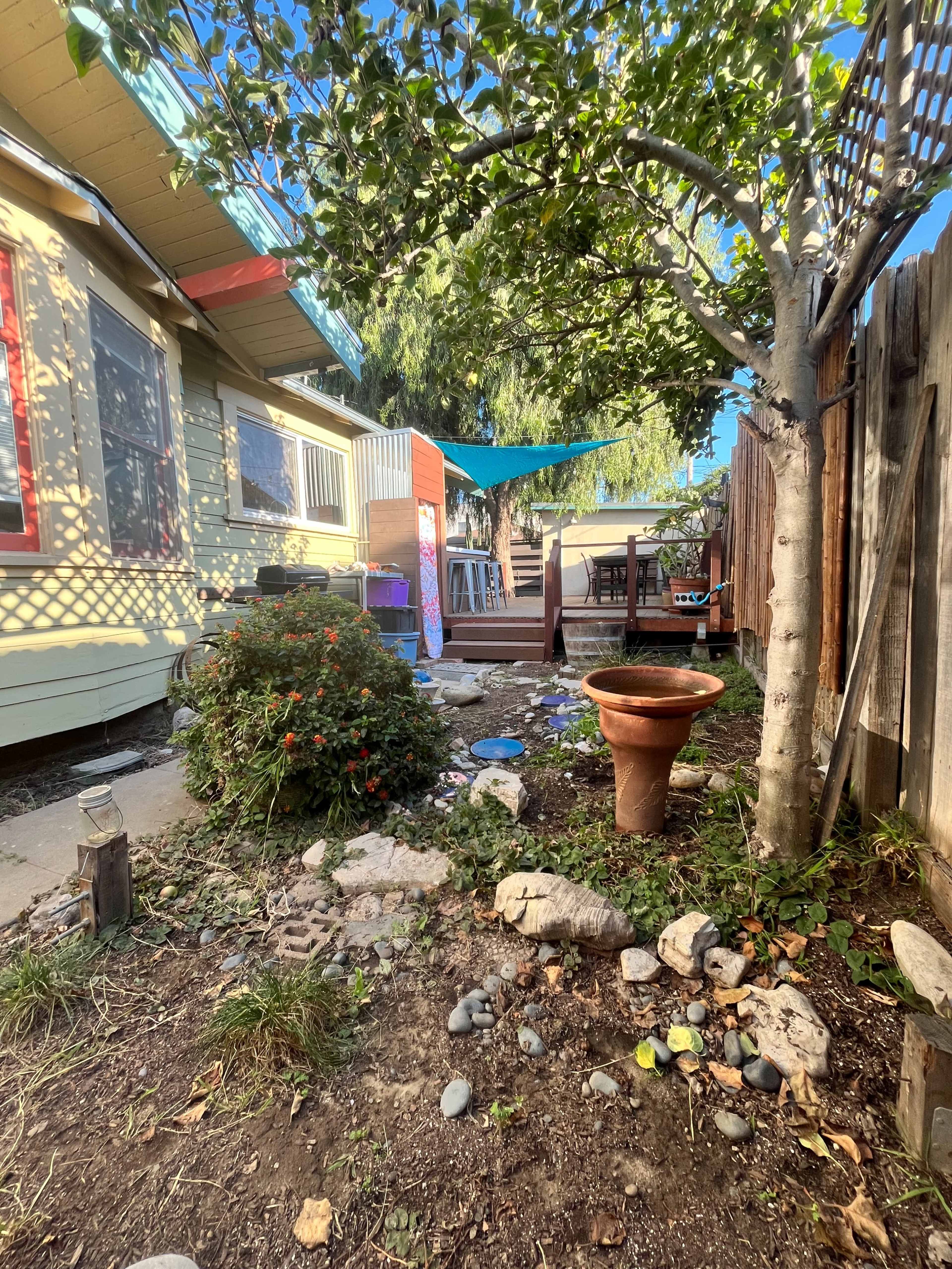 A garden scene features a pathway leading to a wooden deck surrounded by various plants and decorative stones, with a tree providing shade.