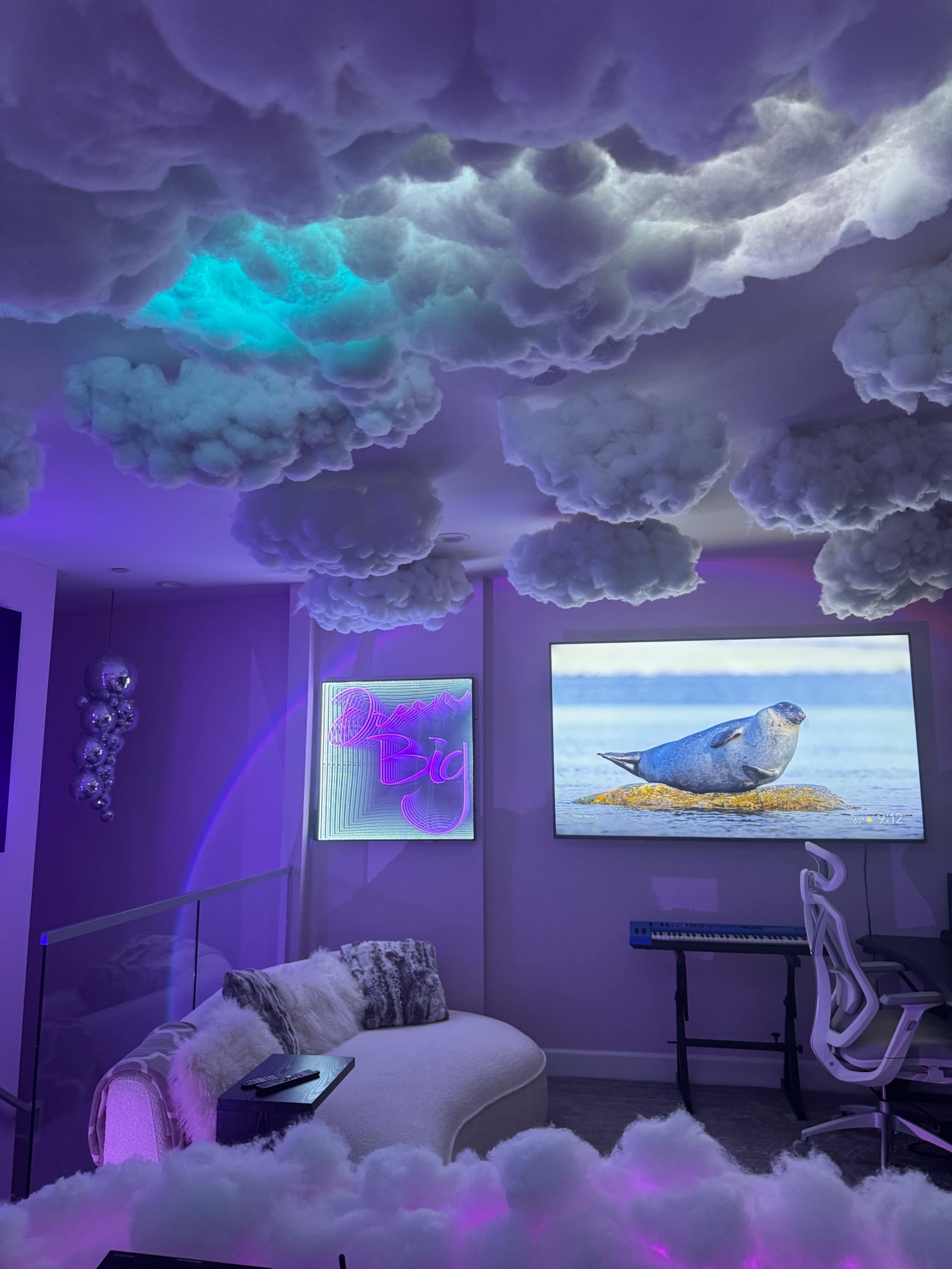 A modern room features a curved white sofa, a large screen displaying a seal, and a ceiling adorned with fluffy cloud-like decorations illuminated by colorful lights.