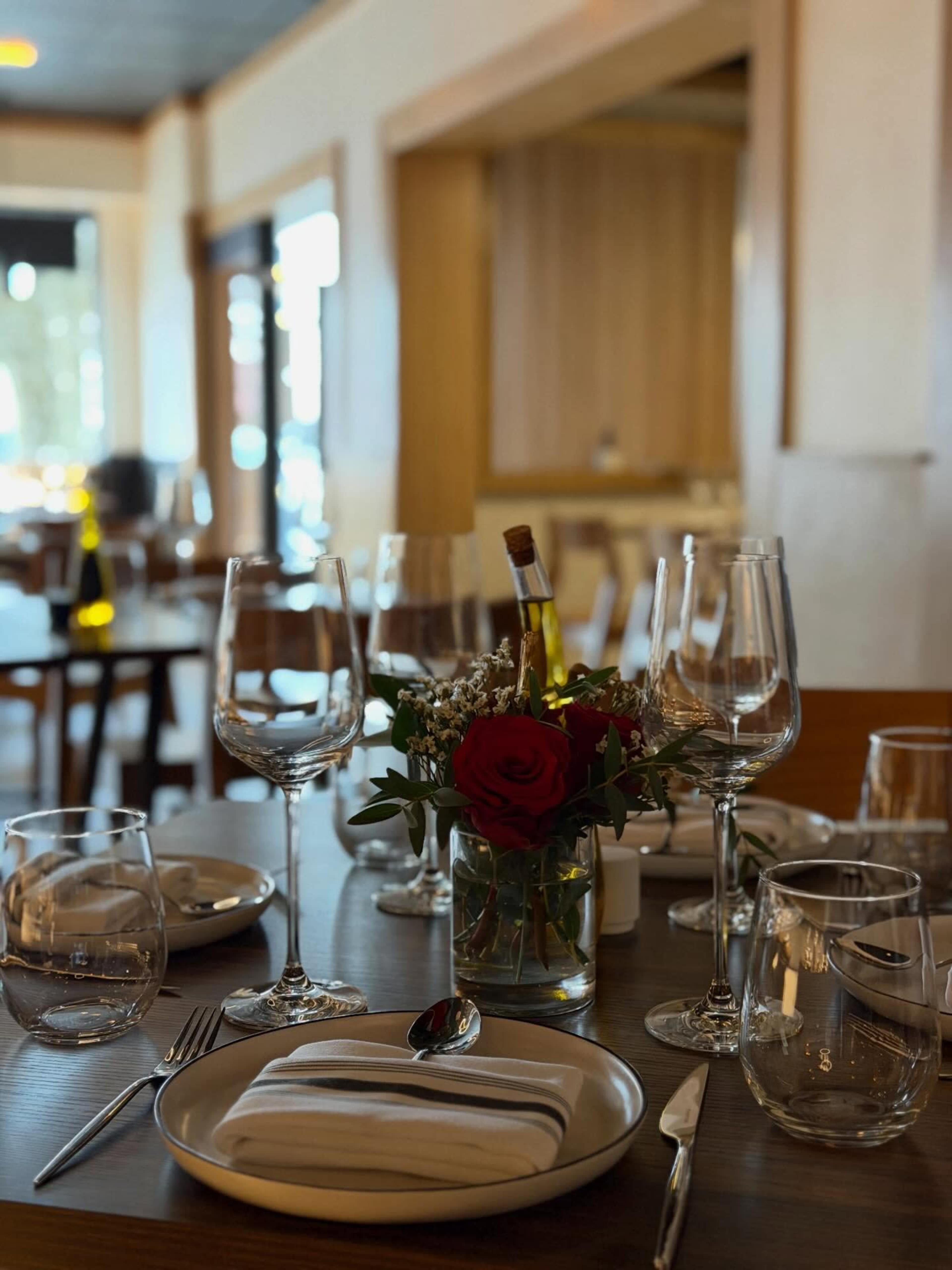 A table is set with elegant glassware, a small vase holding a rose and flowers, and neatly arranged utensils in a restaurant setting.