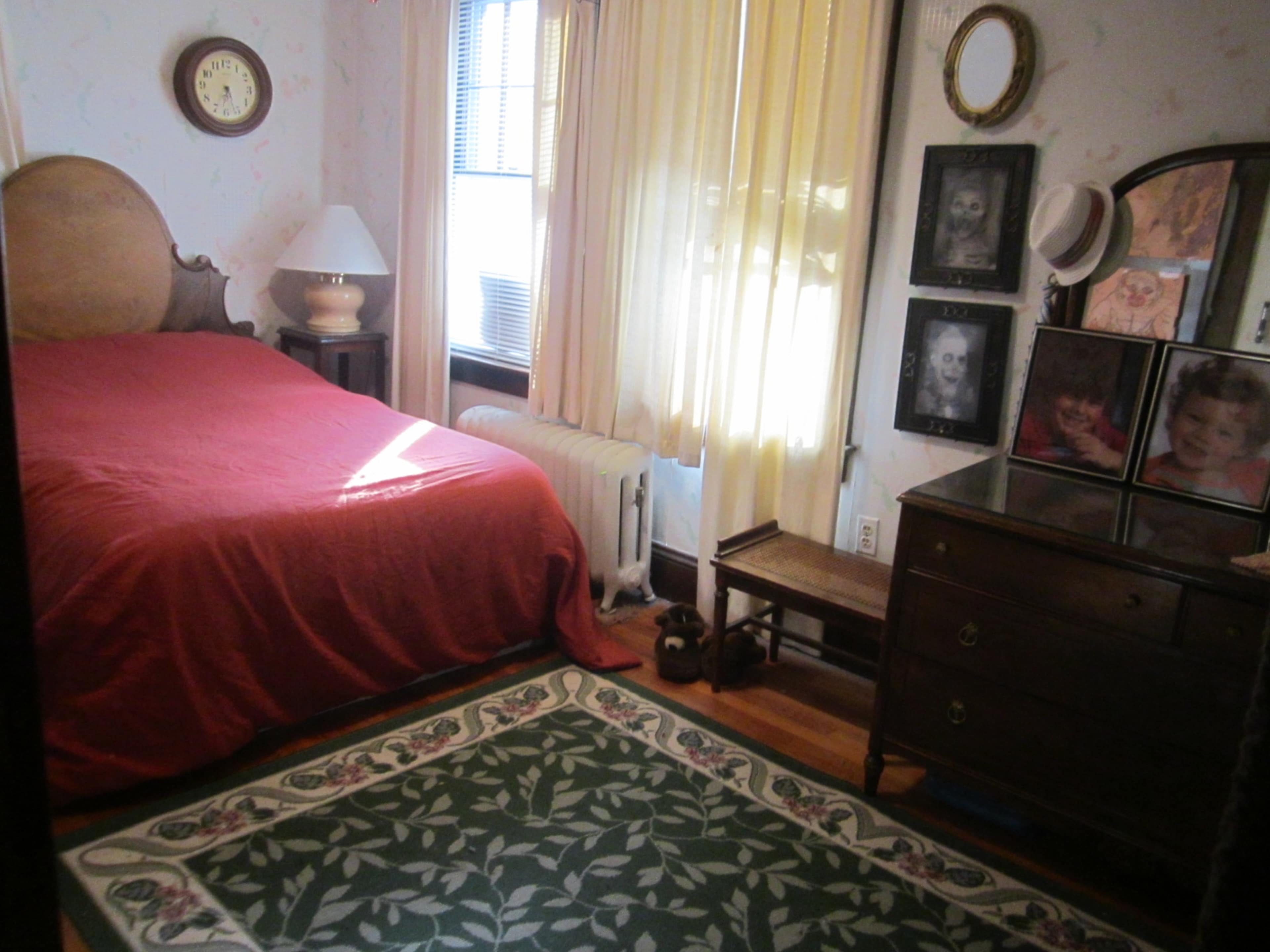 The image shows a bedroom with a red bedspread, a lamp, a window covered by yellow curtains, and a wall adorned with framed photographs.
