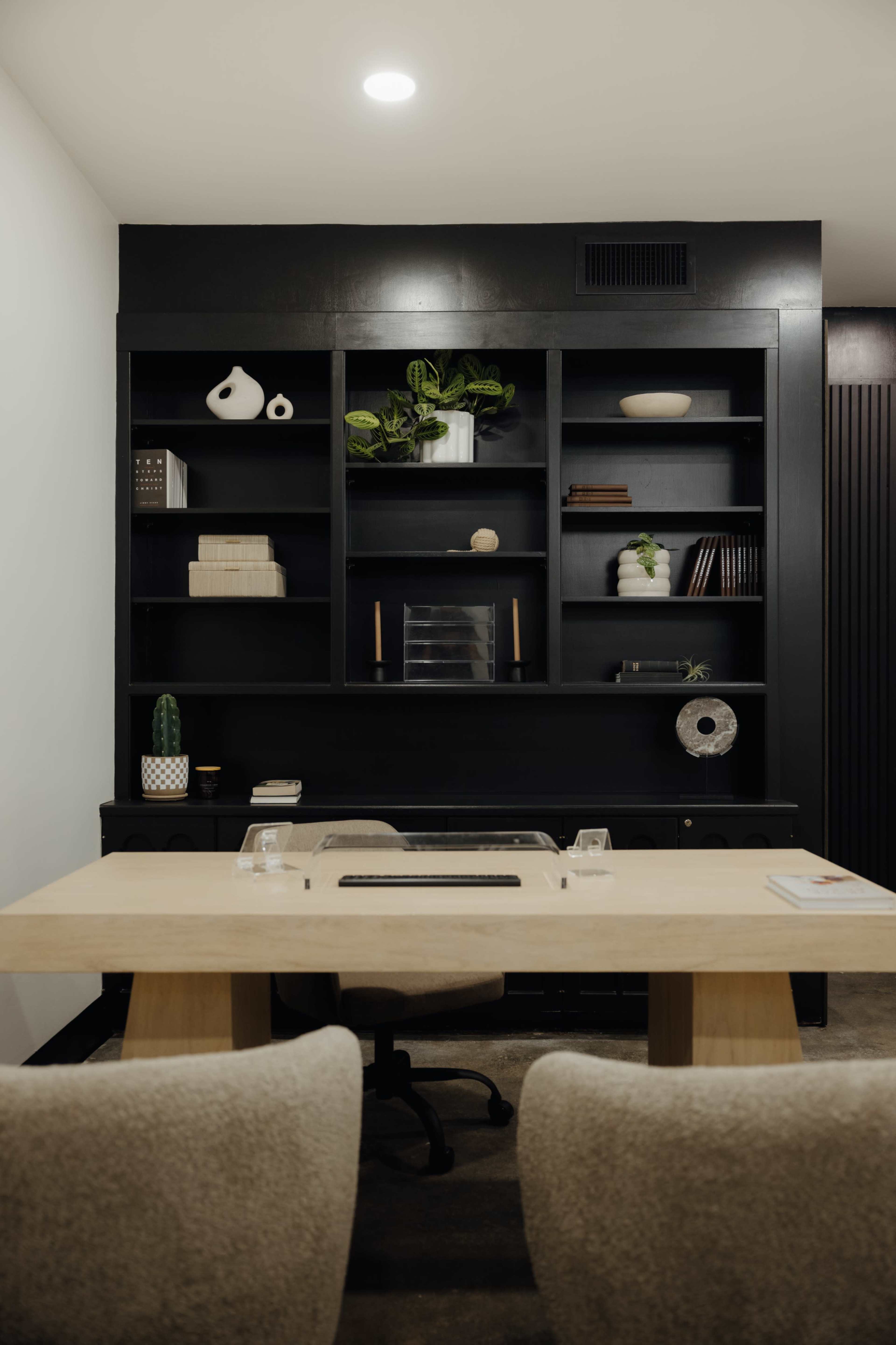 A modern office space features a wooden desk in front of a black bookshelf filled with decorative items and plants.