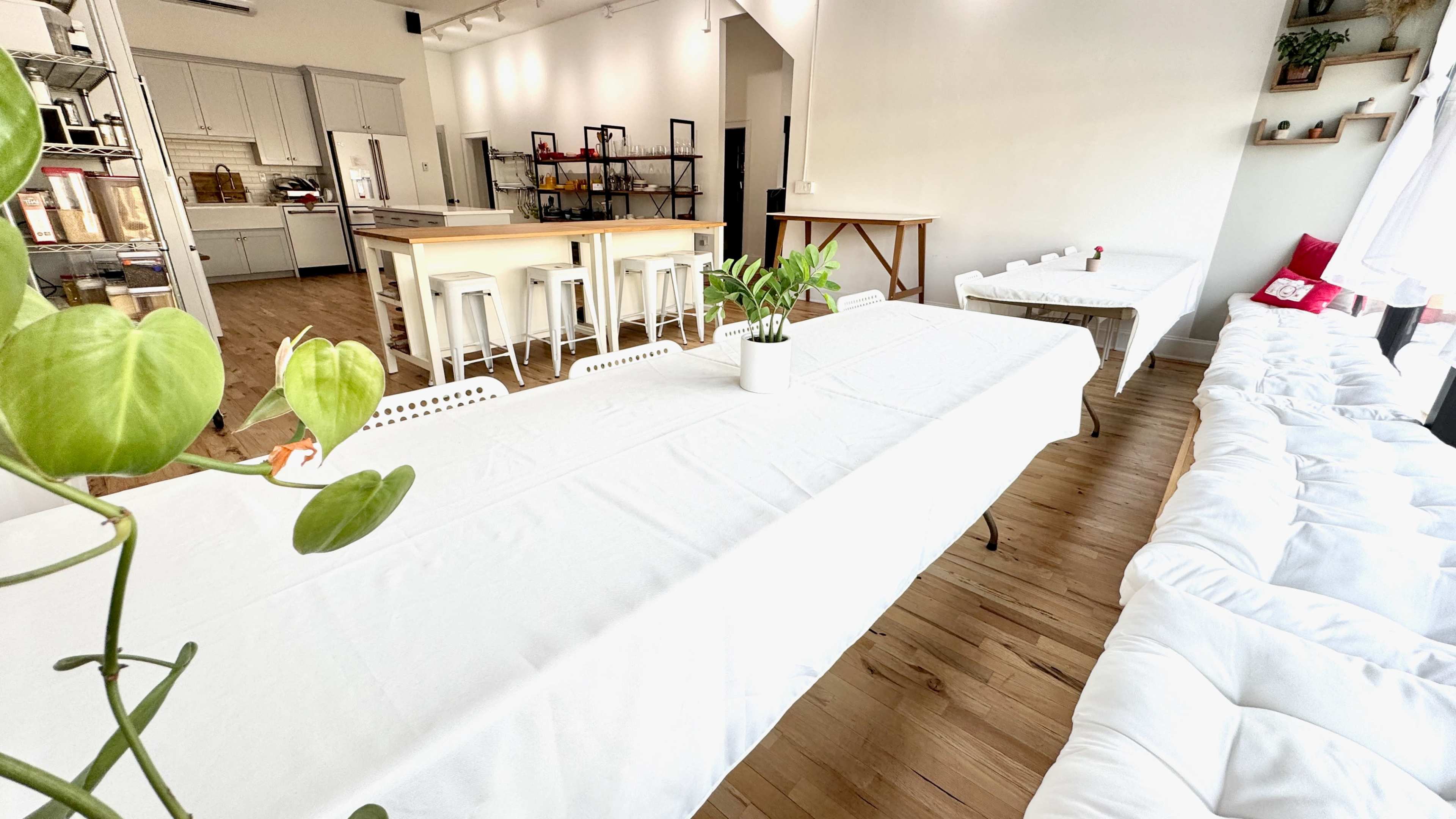 The image shows a bright, minimalist interior space featuring a long white table with matching chairs, a kitchen area in the background, and a cozy seating area along the wall.
