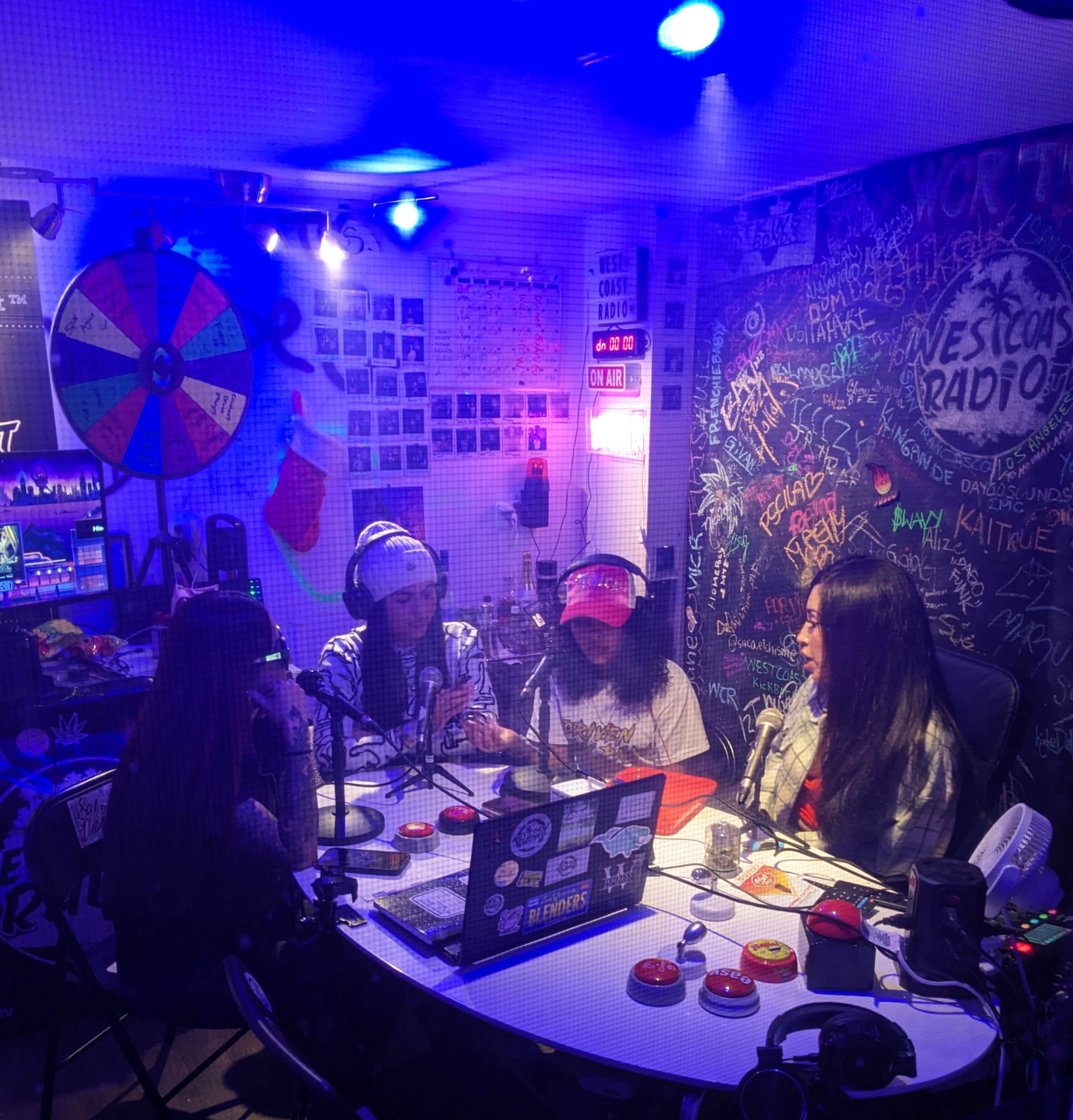 Four people are seated at a table in a radio studio, surrounded by colorful decorations and equipment, while discussing into microphones.