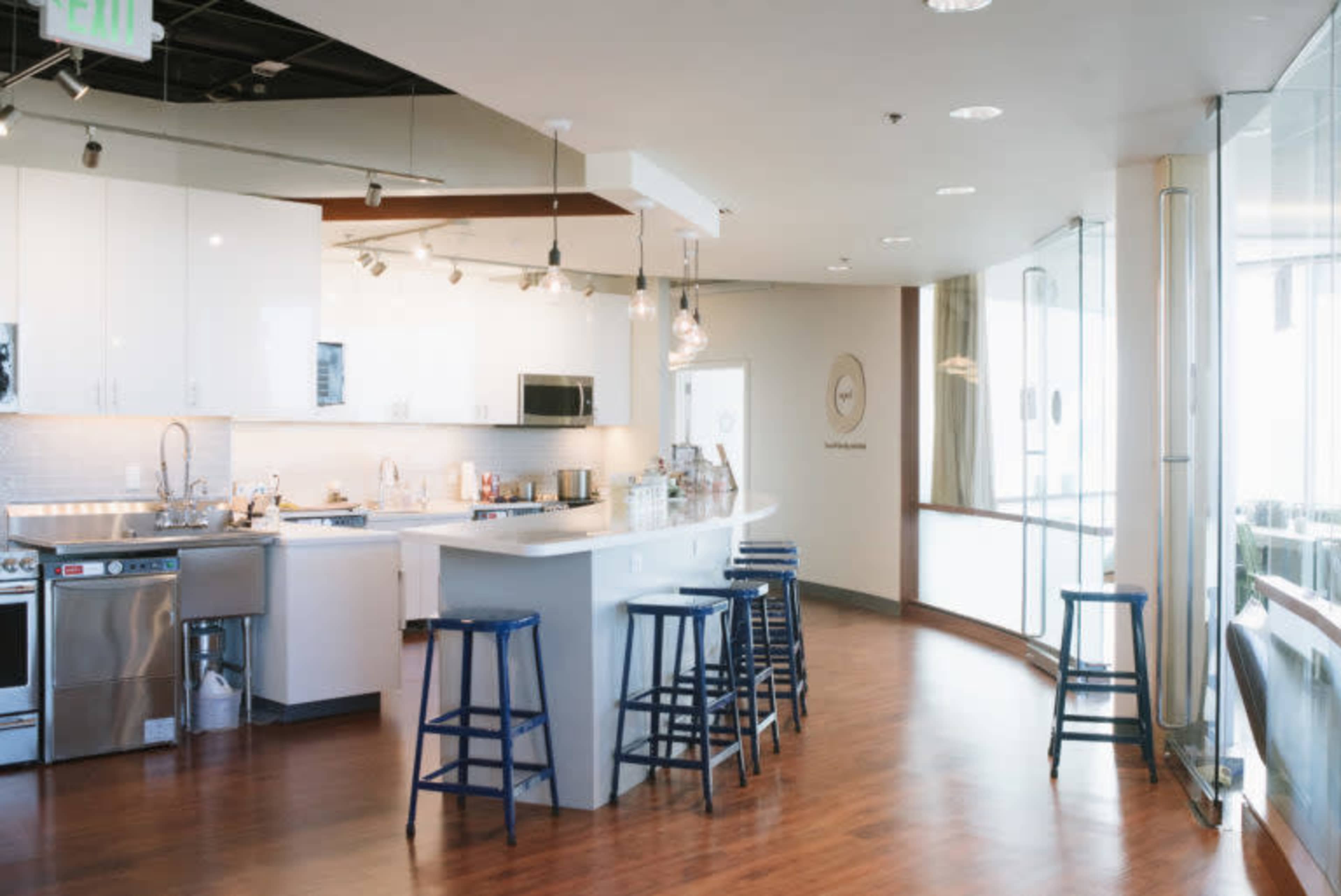 The image shows a modern kitchen area with white cabinetry, stainless steel appliances, and bar stools along a white countertop.
