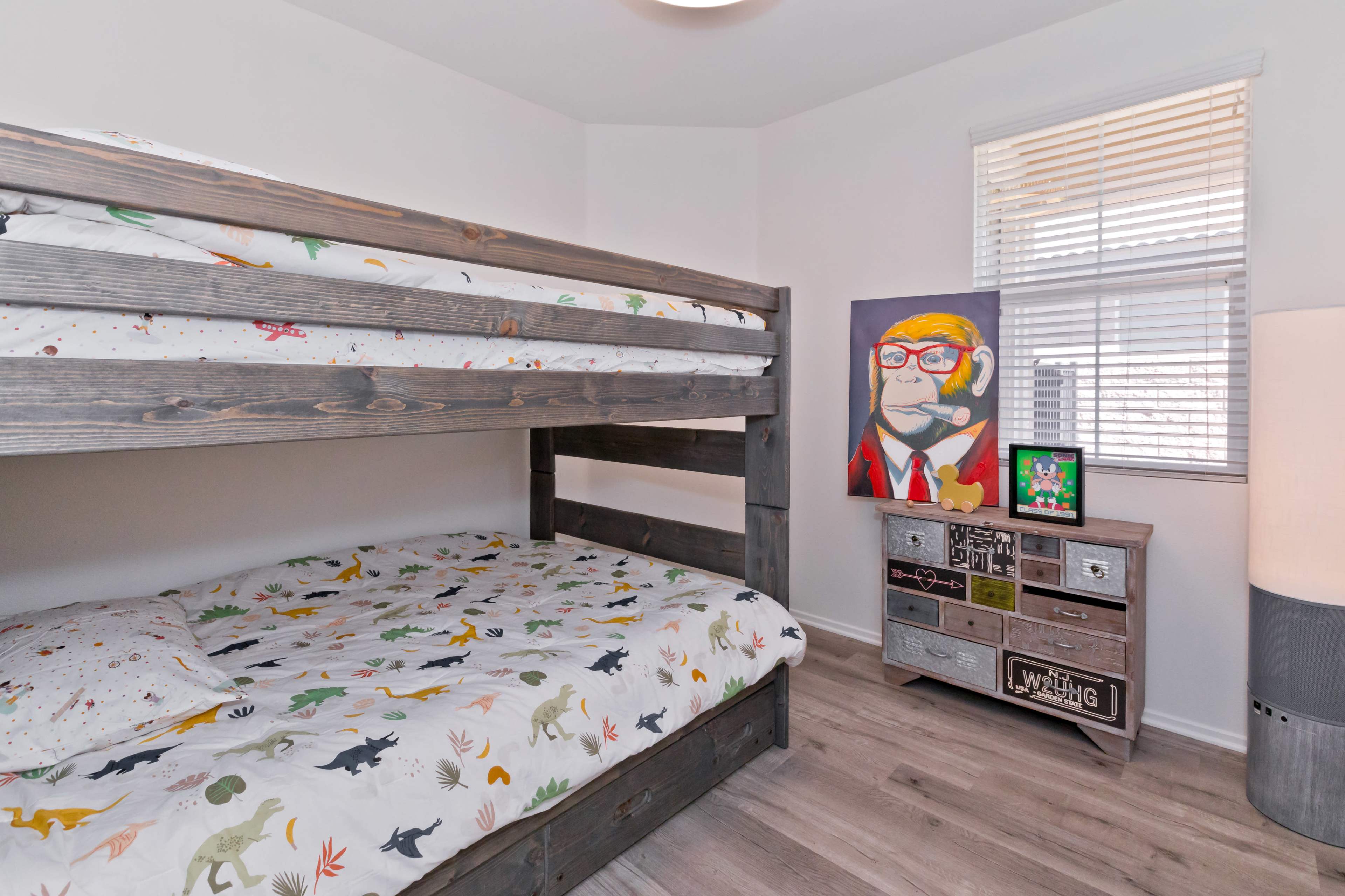 A bunk bed with colorful dinosaur-patterned bedding is positioned in a brightly lit room, featuring a large painting and a wooden dresser.