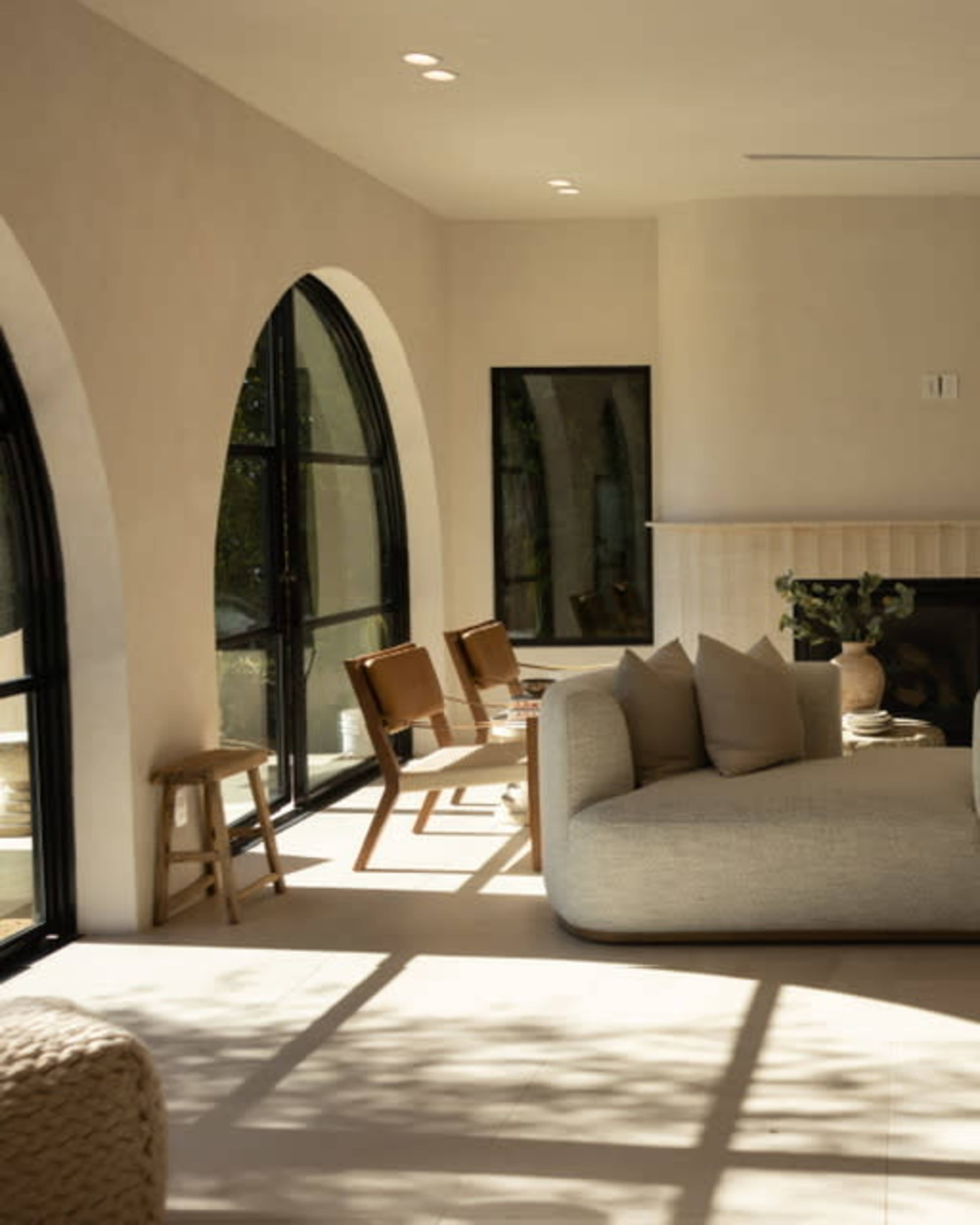 The image shows a modern interior space with large arched windows, a simple white sofa, wooden chairs, and a fireplace.
