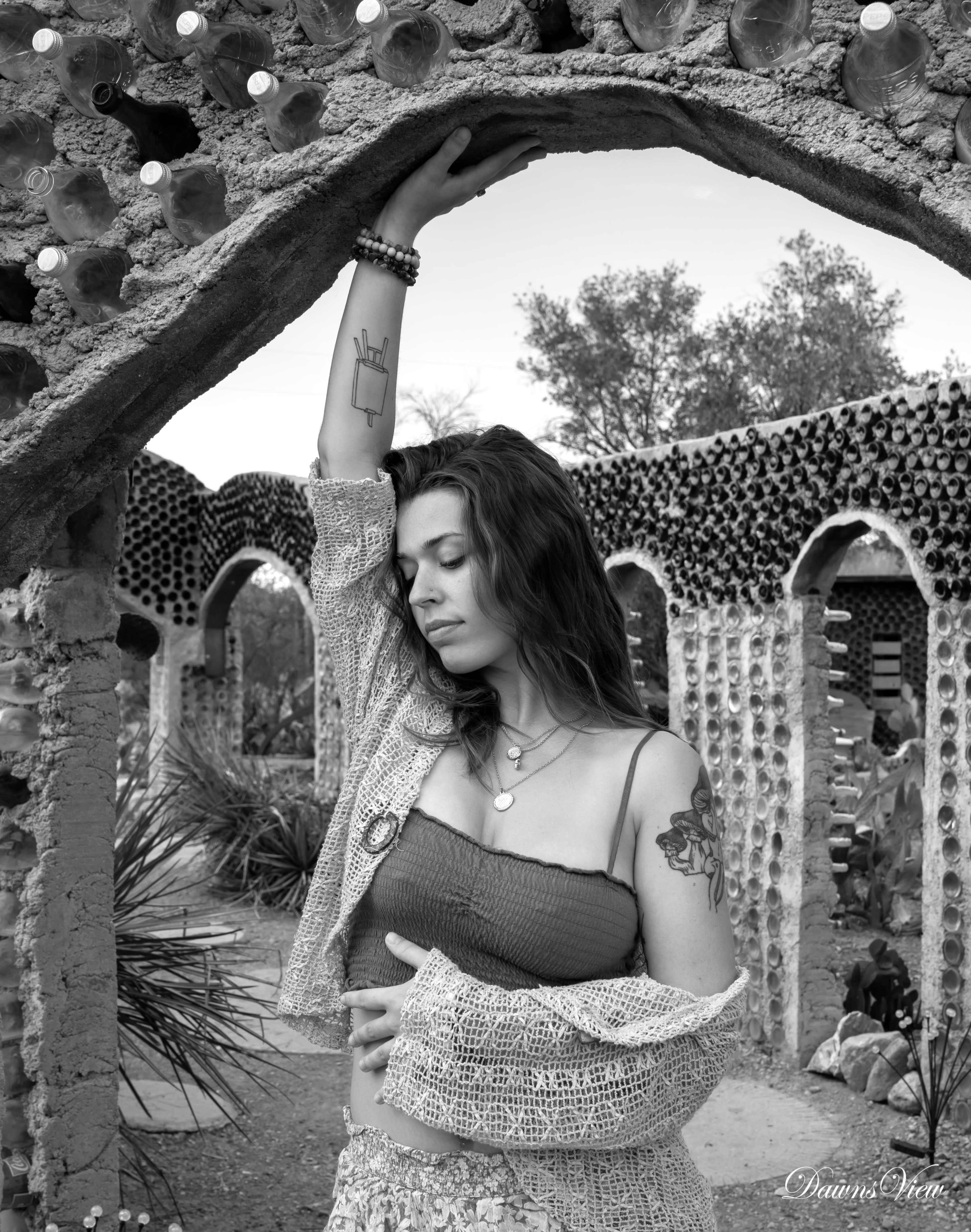 A woman in a patterned outfit poses under an archway made of bottles in a desert-like setting.