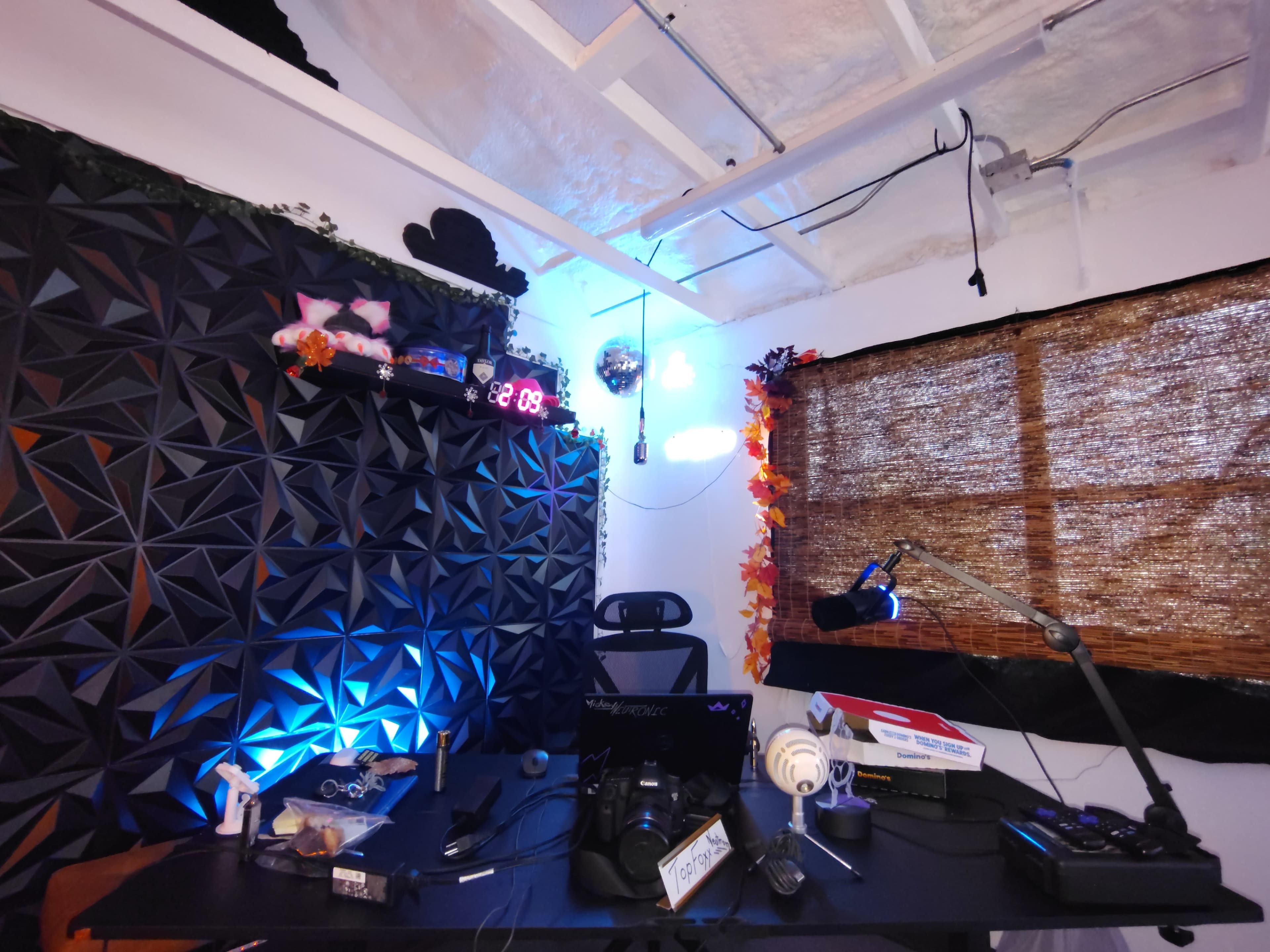 The image shows a small, modern home office featuring a black wall panel with geometric designs, a desk with various electronic devices, and a window partially covered by a woven bamboo shade.