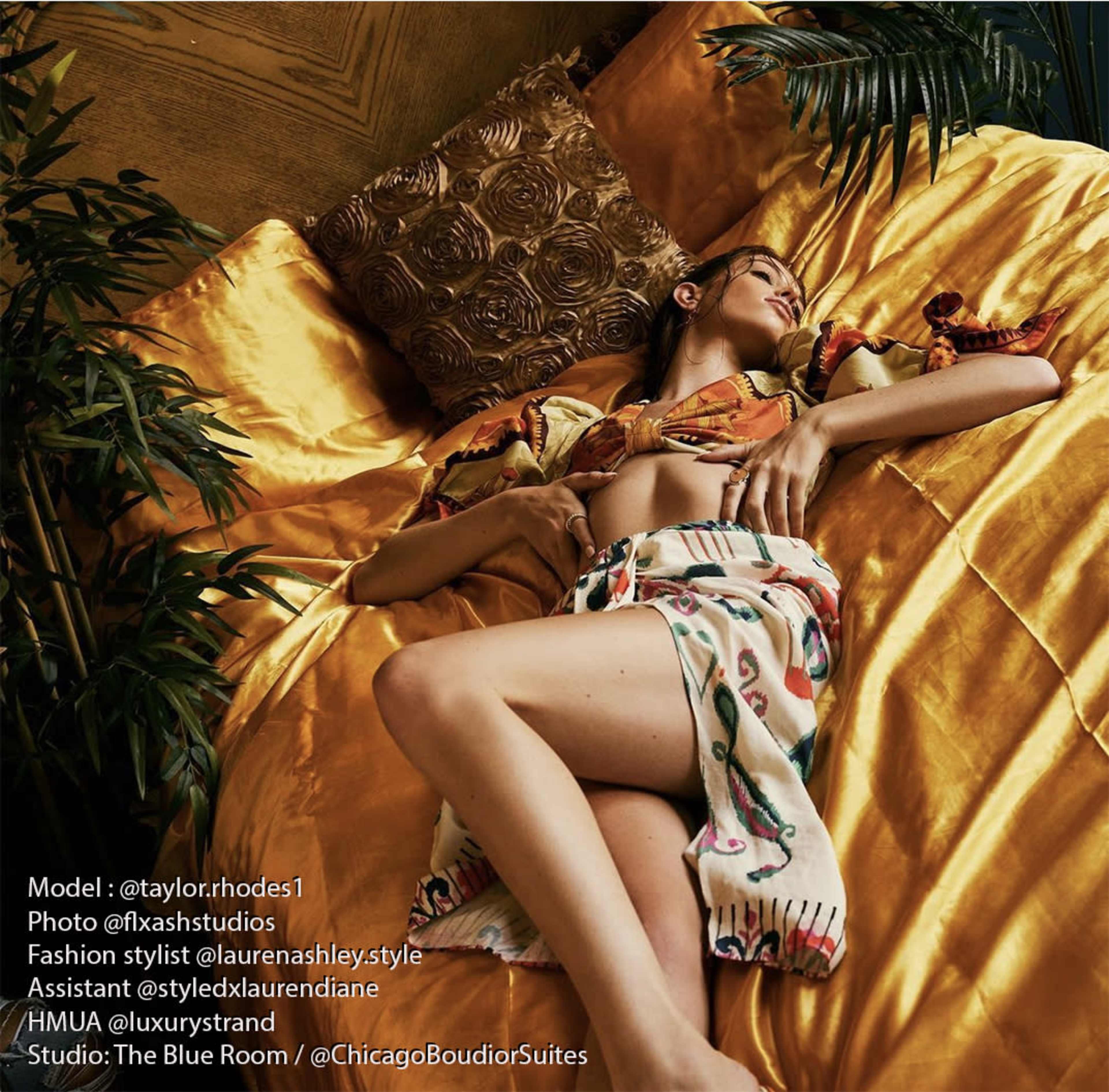 A woman in a colorful outfit is lying on a golden bedspread in a stylish room.