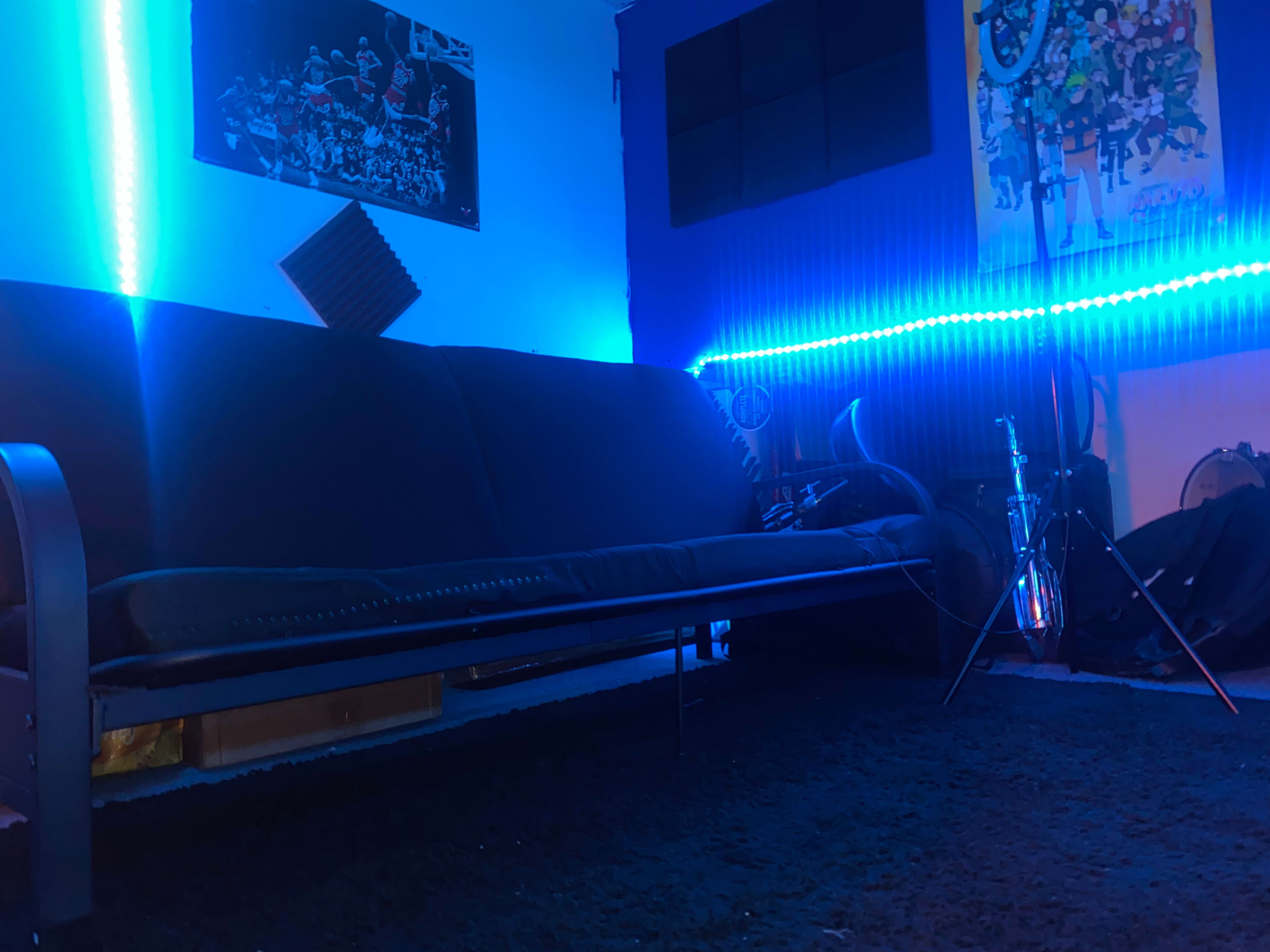 A dimly lit room featuring a black sofa, blue LED lights along the walls, and musical instruments in the corner.