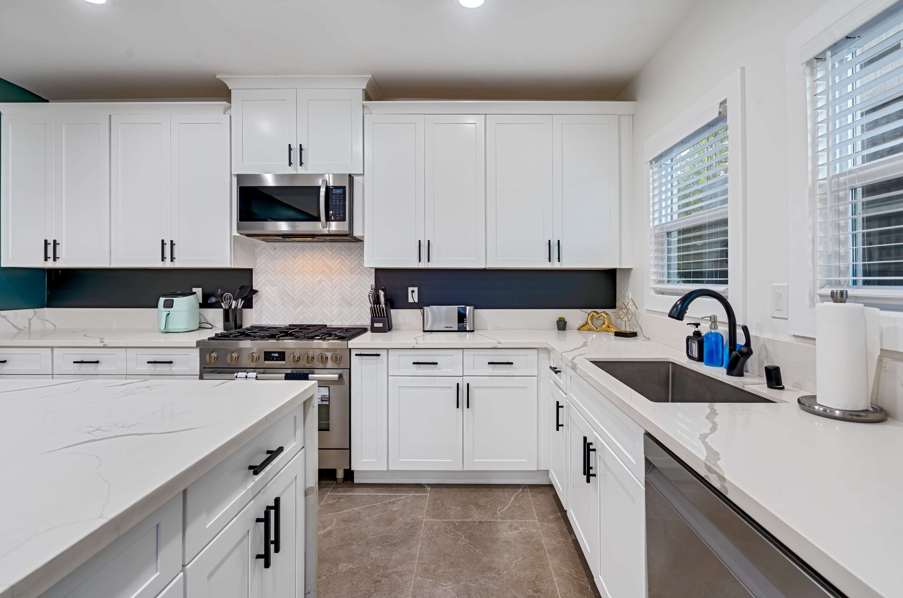The image shows a modern kitchen featuring white cabinets, stainless steel appliances, and a marble countertop, with a sink and a window providing natural light.