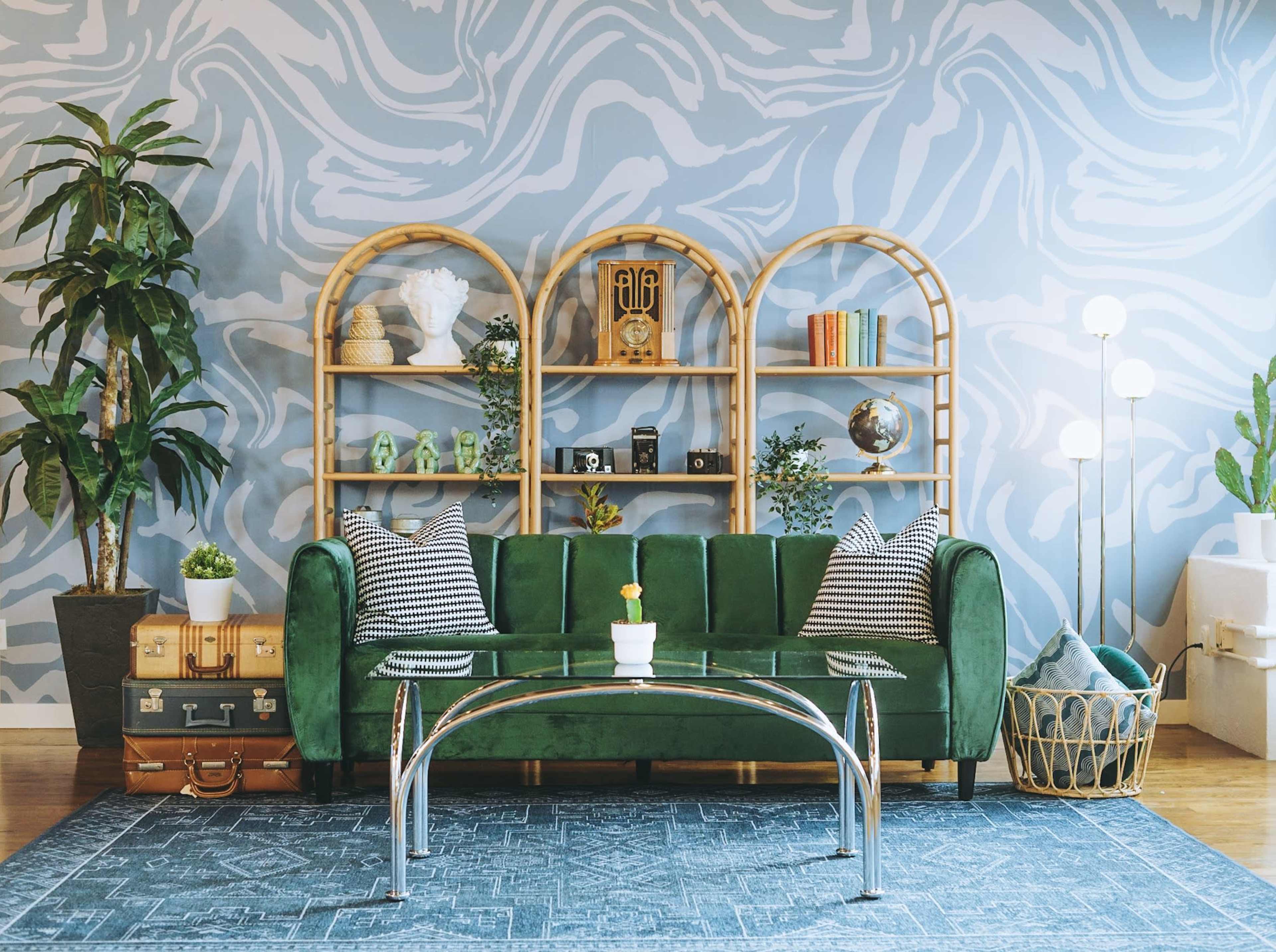A green velvet sofa is positioned in front of a light blue marbled wall, accompanied by a glass coffee table and decorated shelves filled with various items, plants, and a patterned area rug.