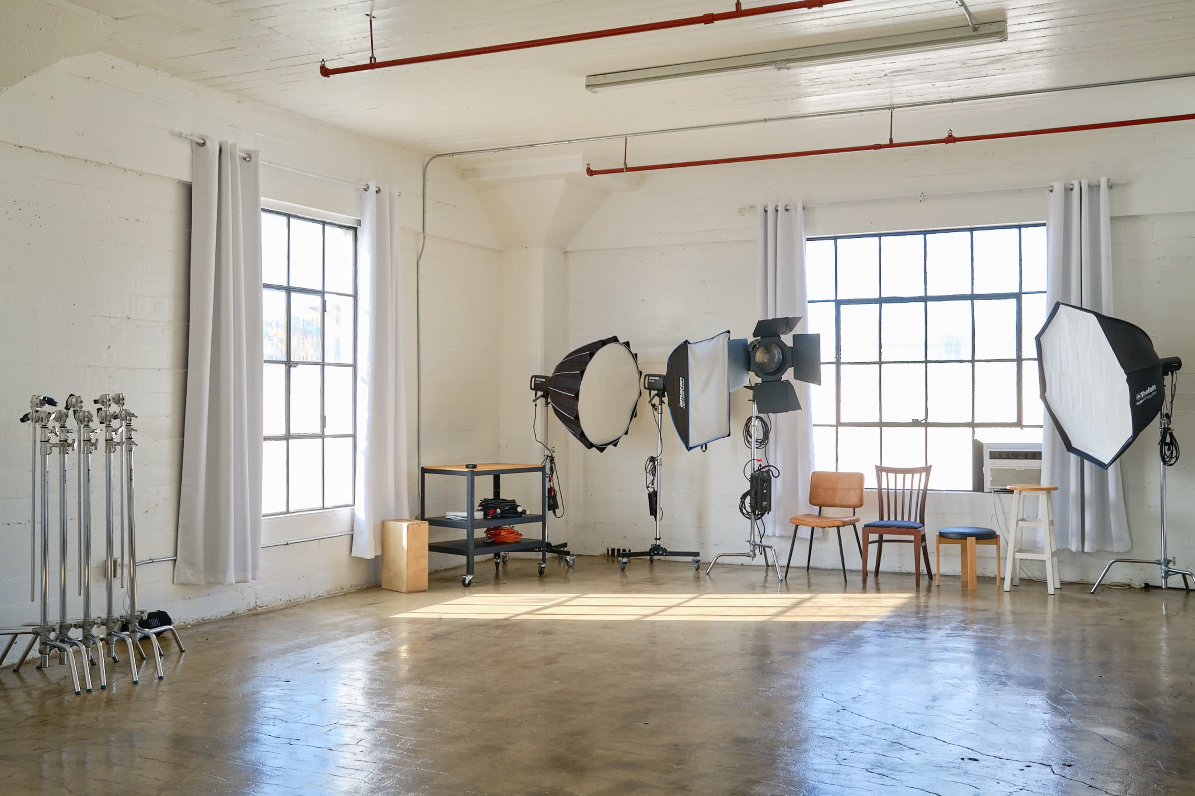 The scene features a spacious photography studio with large windows, softbox lights, and a few chairs along with studio equipment.