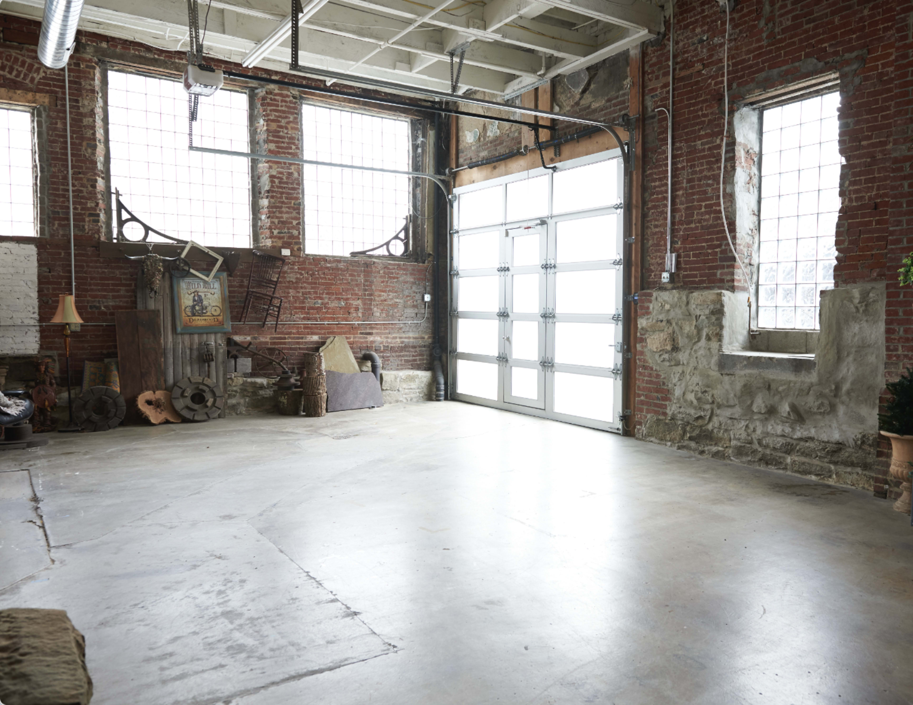The image shows an empty industrial space with exposed brick walls, large windows, and concrete flooring.