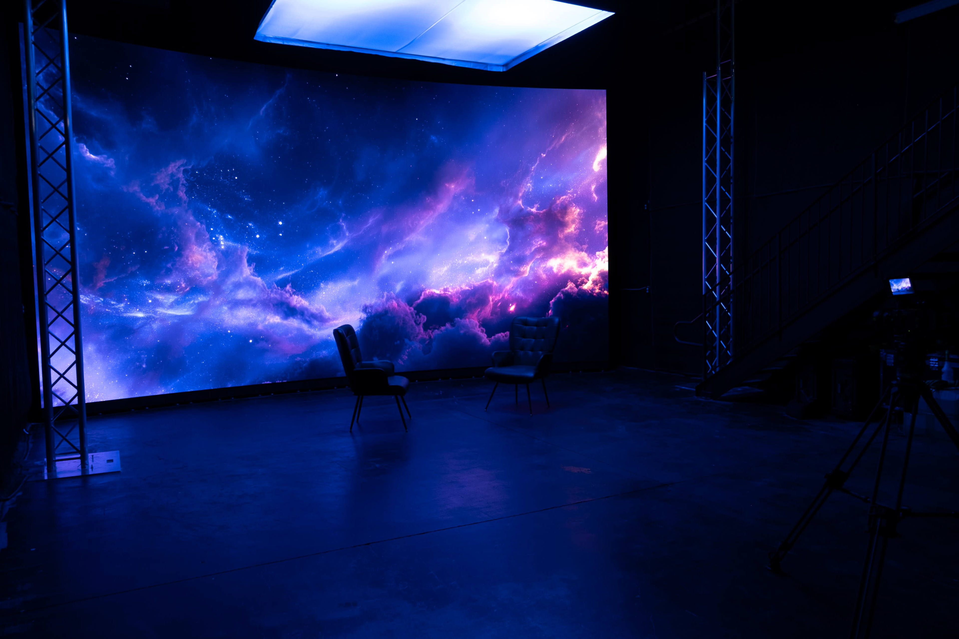 The image shows a studio with two chairs positioned in front of a large, luminous backdrop featuring a colorful cosmic scene.