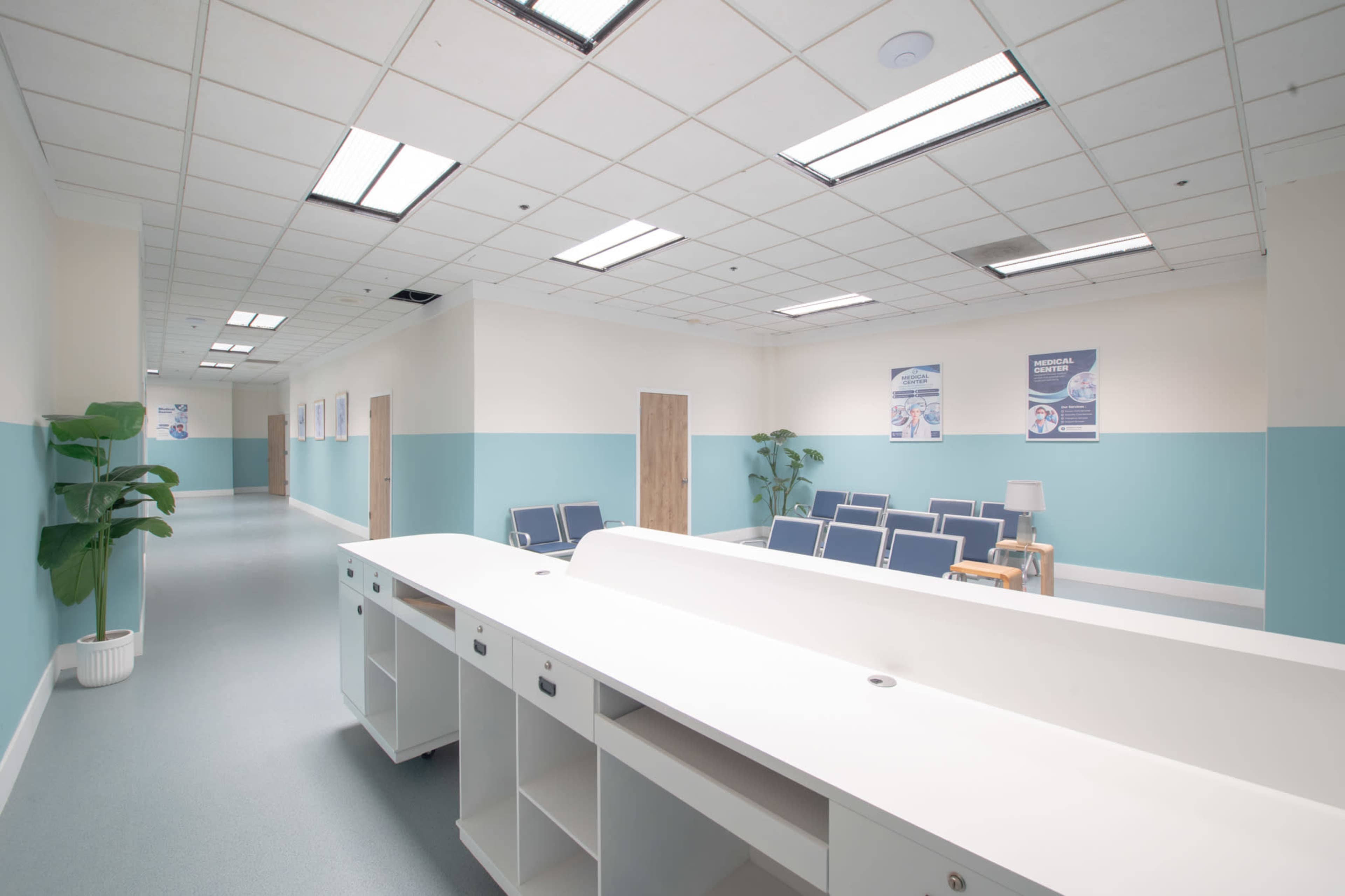 The image shows a brightly lit medical waiting area with teal walls, a reception desk, and rows of blue chairs.