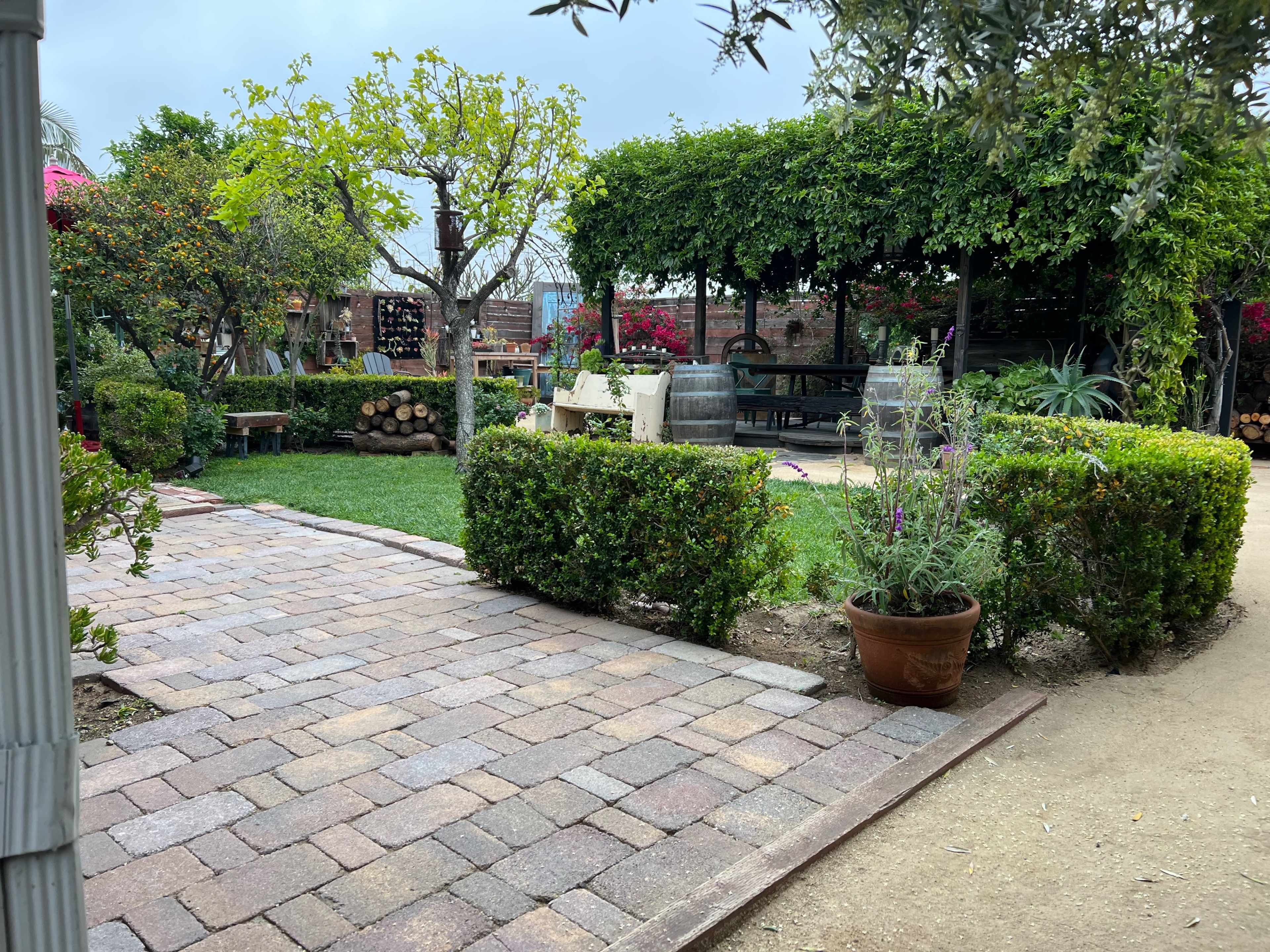 A brick pathway leads through a garden with lush greenery, potted plants, and shaded seating areas.