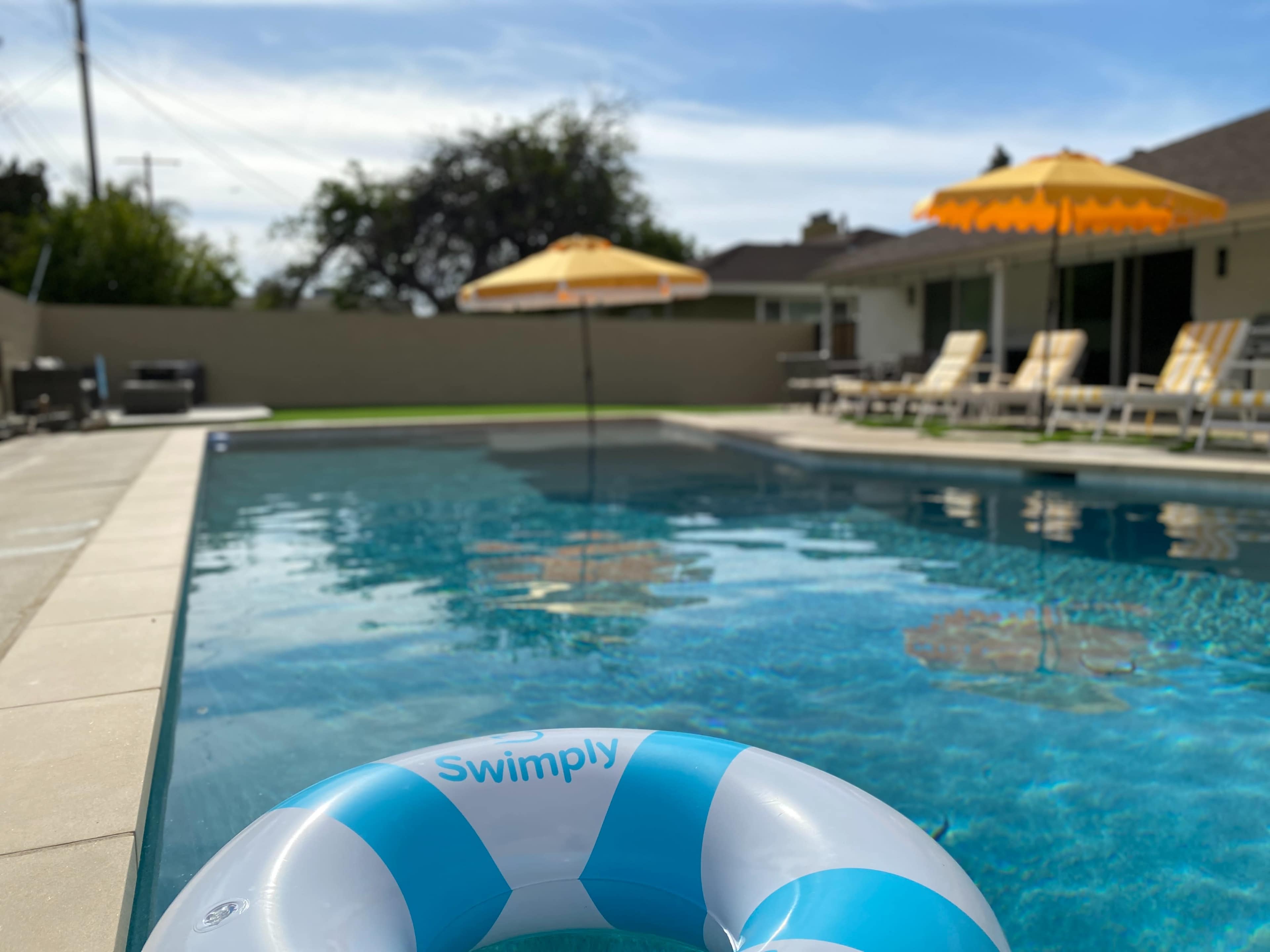 A swimming pool with a blue and white flotation ring, sun umbrellas, and lounge chairs is set in a residential backyard.