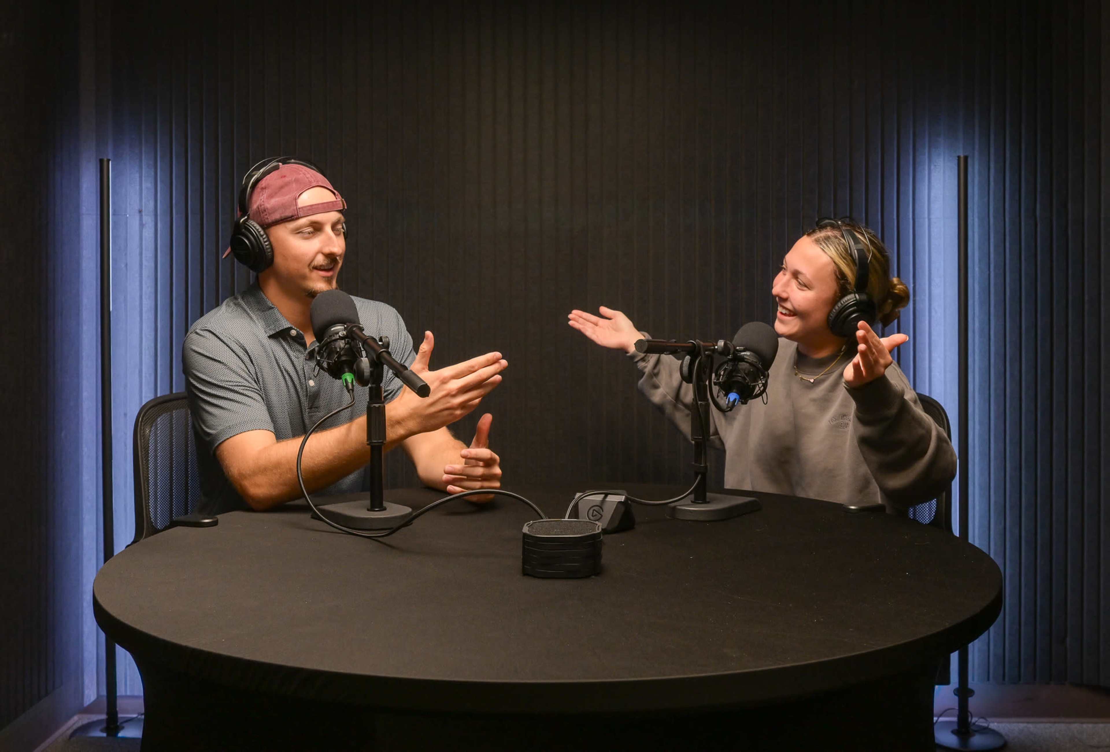 A man and a woman are sitting at a round table in a podcast studio, both wearing headphones and speaking animatedly into microphones.