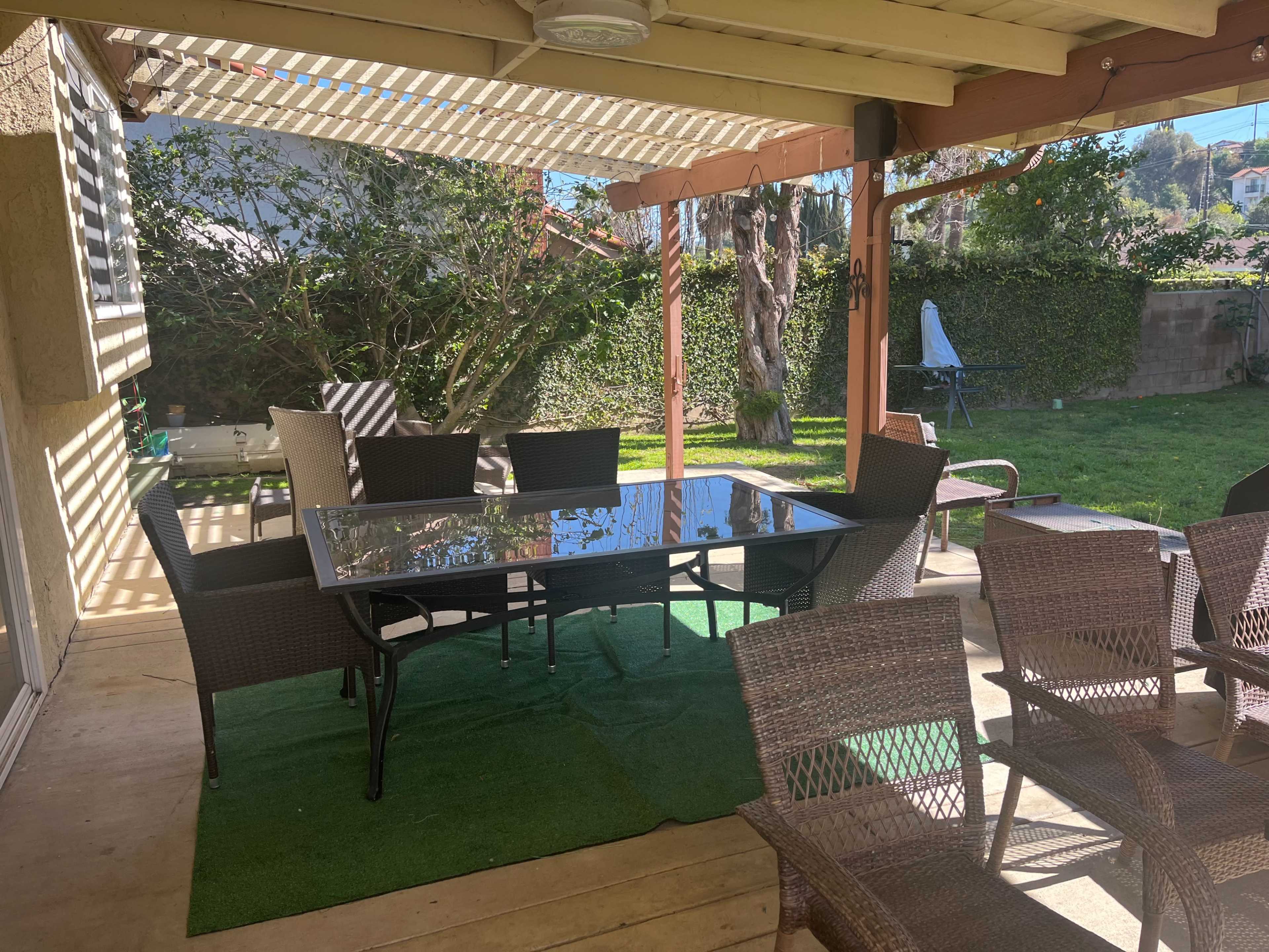 A covered patio features a glass dining table surrounded by several wicker chairs, with a grassy area and plants visible in the background.