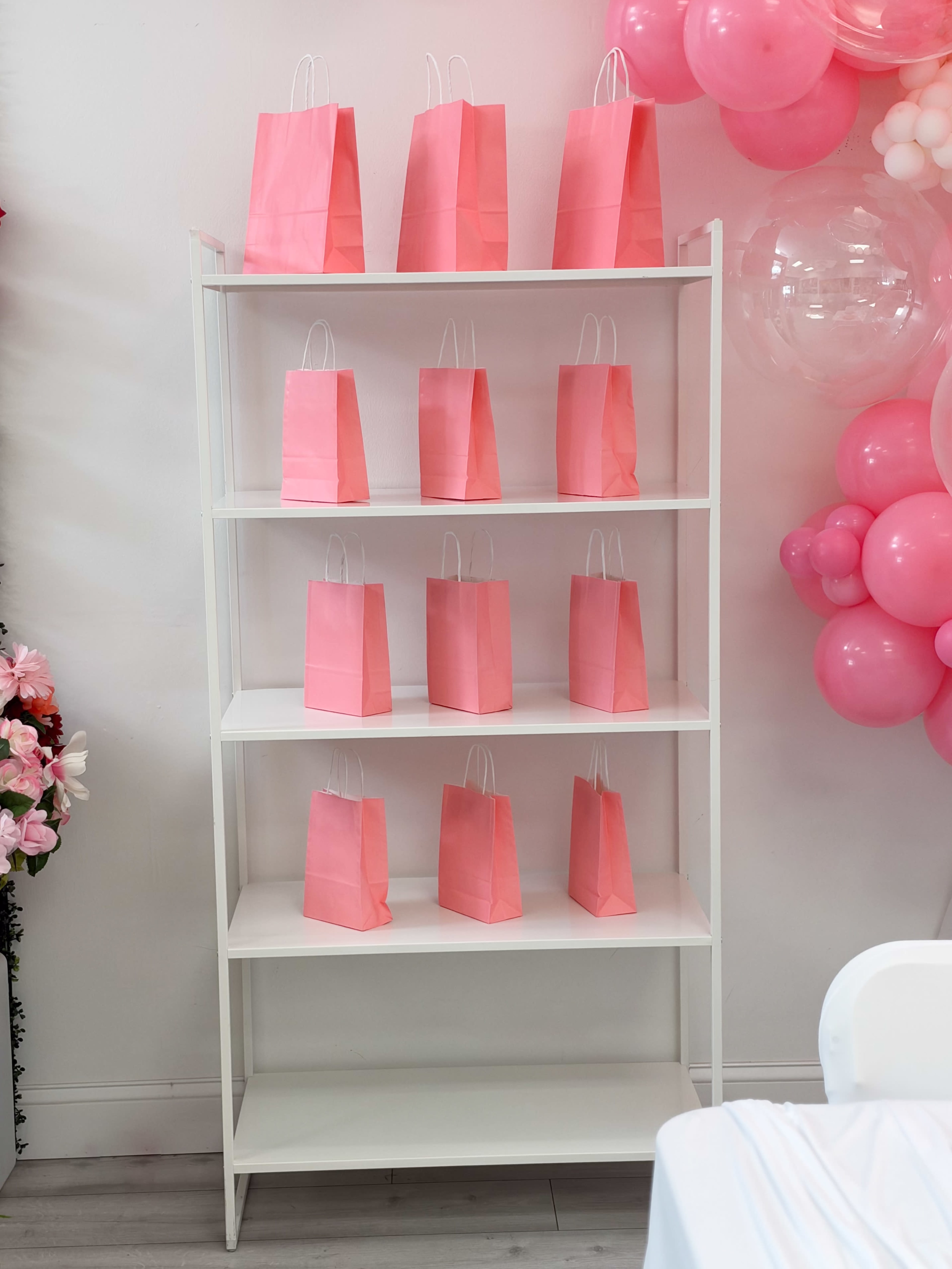 A white shelving unit displays several pink gift bags organized on its shelves.